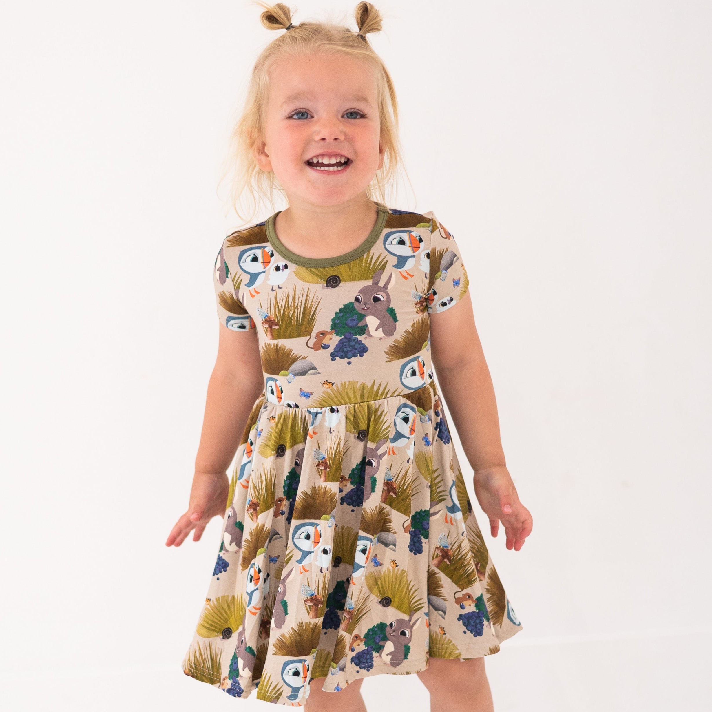A young girl smiles in a tan dress with a Puffin Rock™ woodland print.