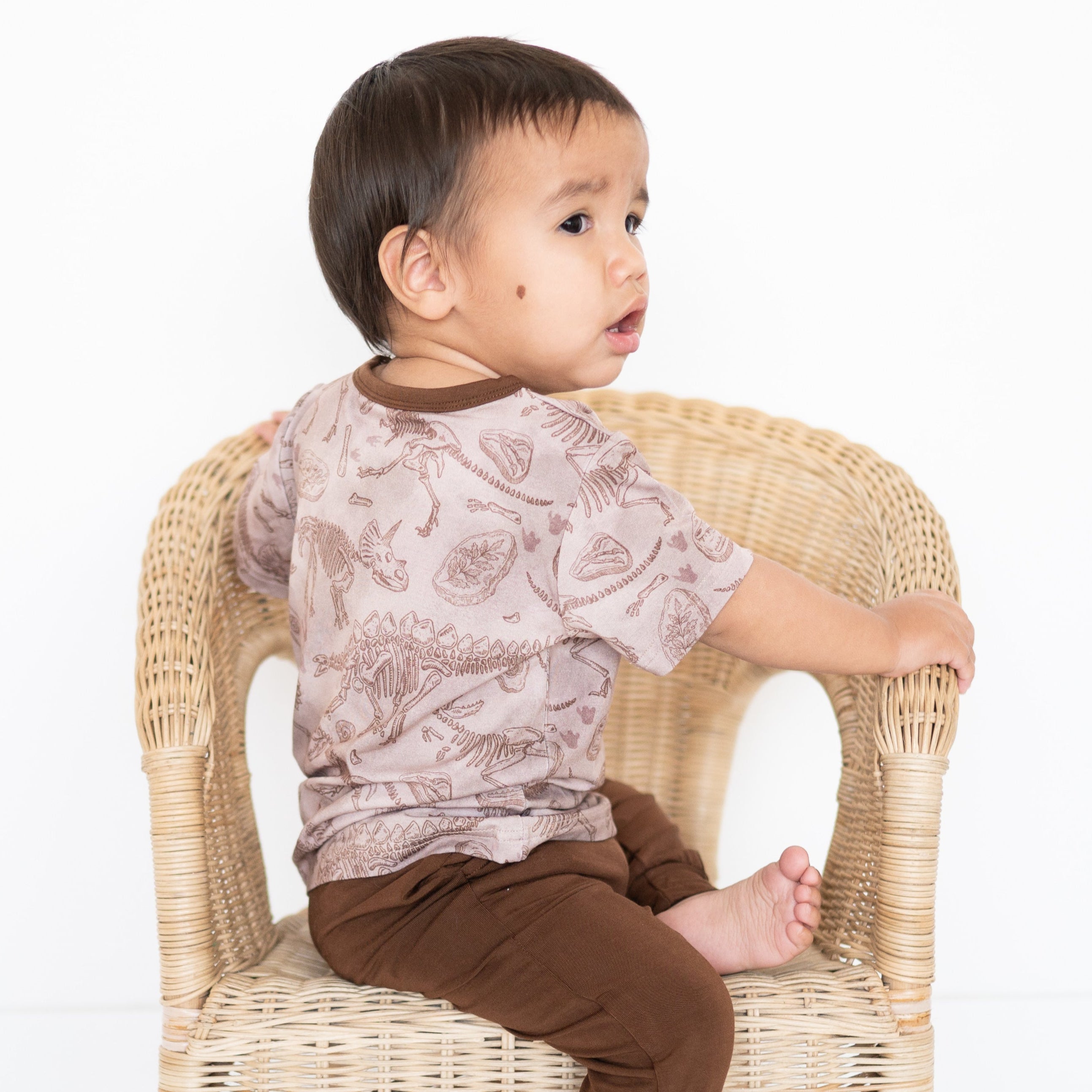 Toddler wearing a light brown t-shirt with a fossil print, sitting on a wicker chair.