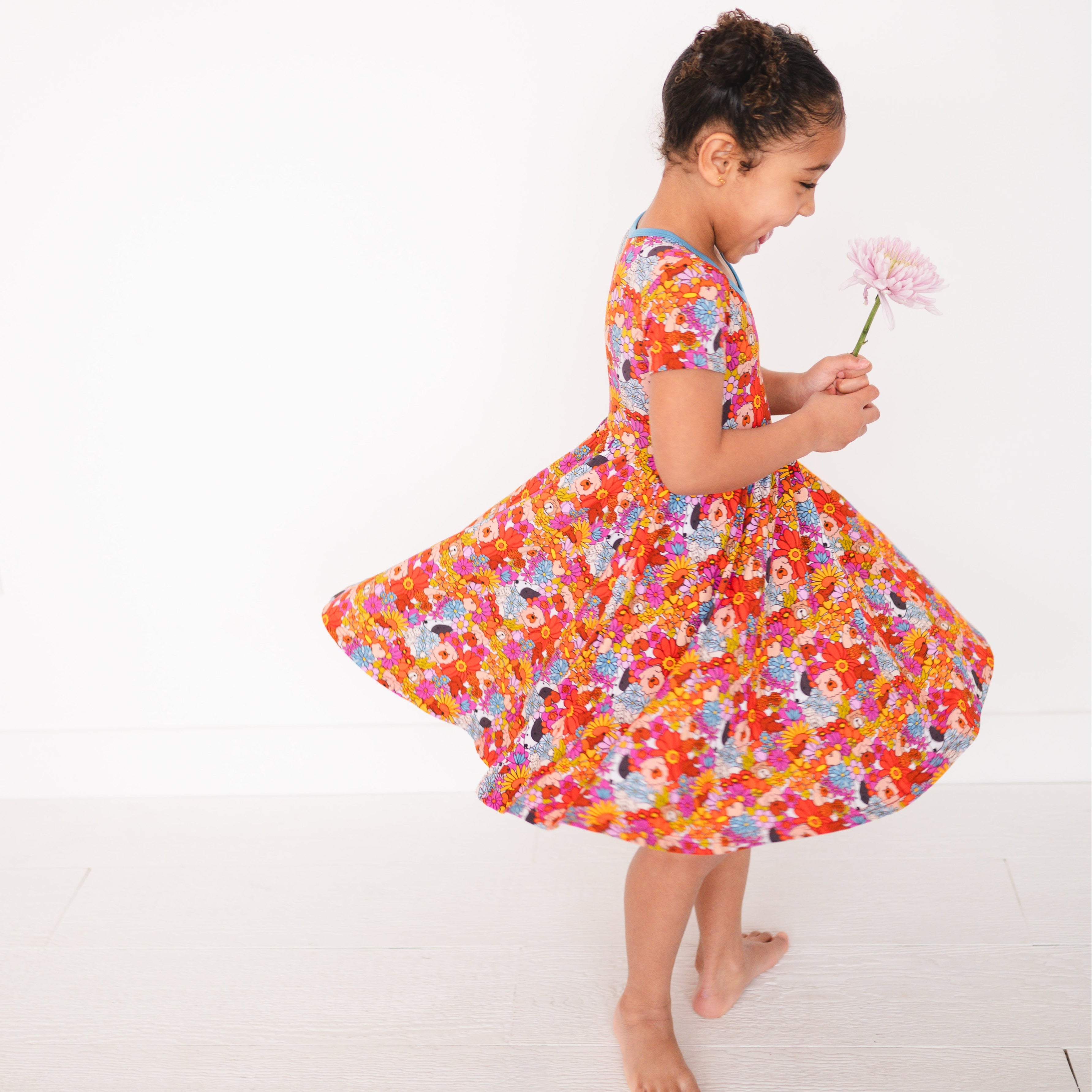 A young girl in a colorful floral dress twirls with a pink flower.