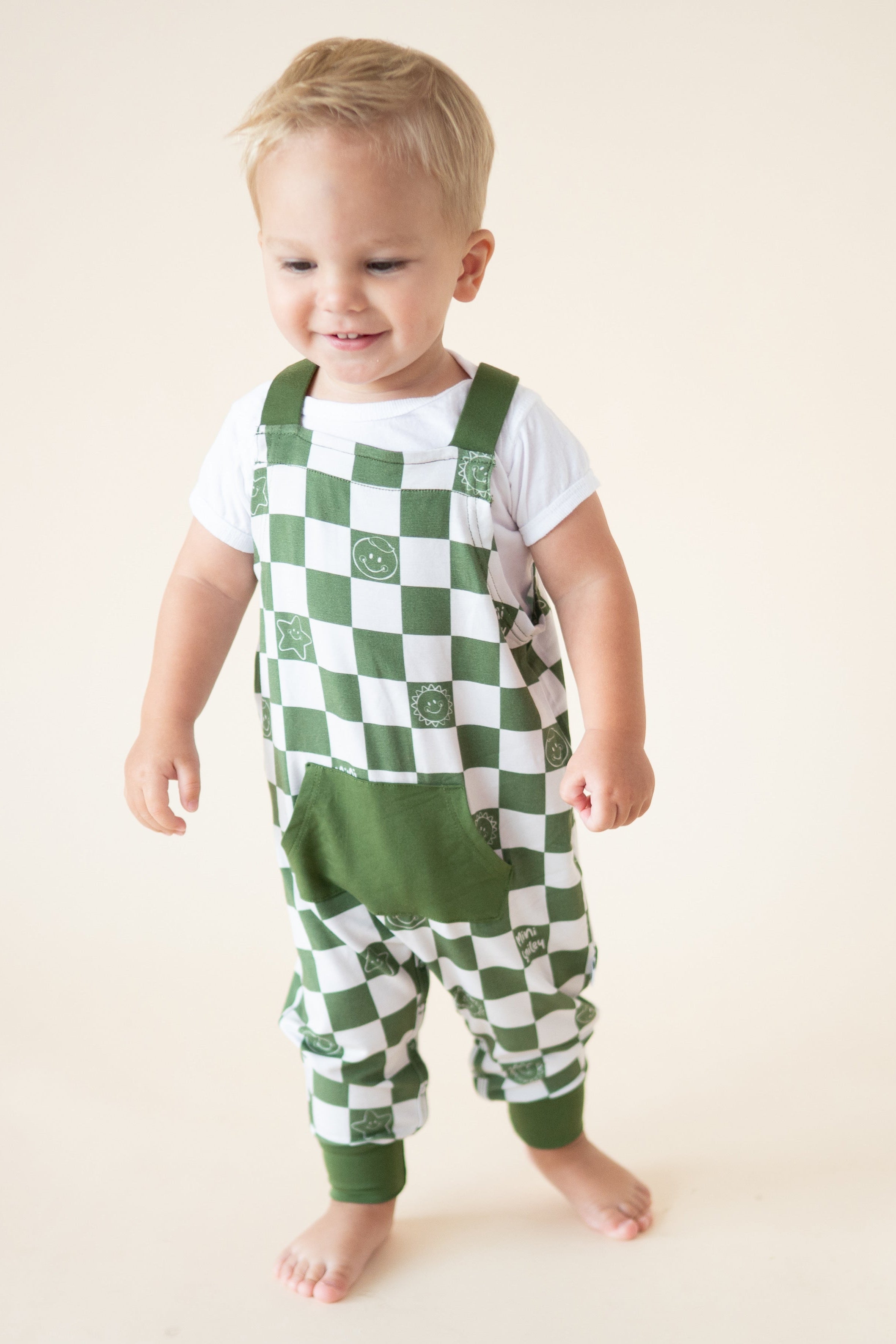 A baby boy wears green and white checkered overalls with smiley face prints over a white t-shirt.