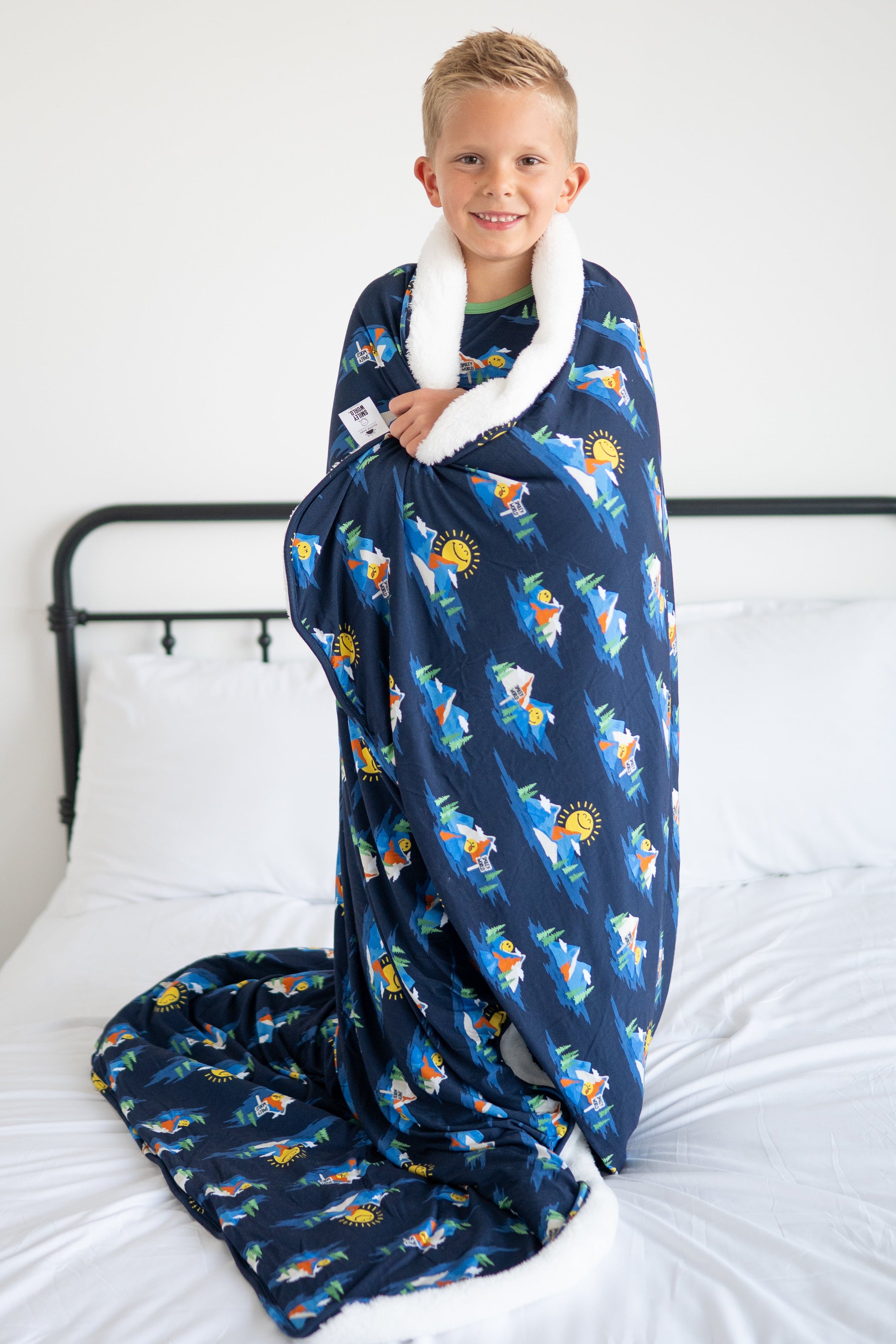 A boy wrapped in a navy blue blanket with a SmileyWorld mountain print and white trim.