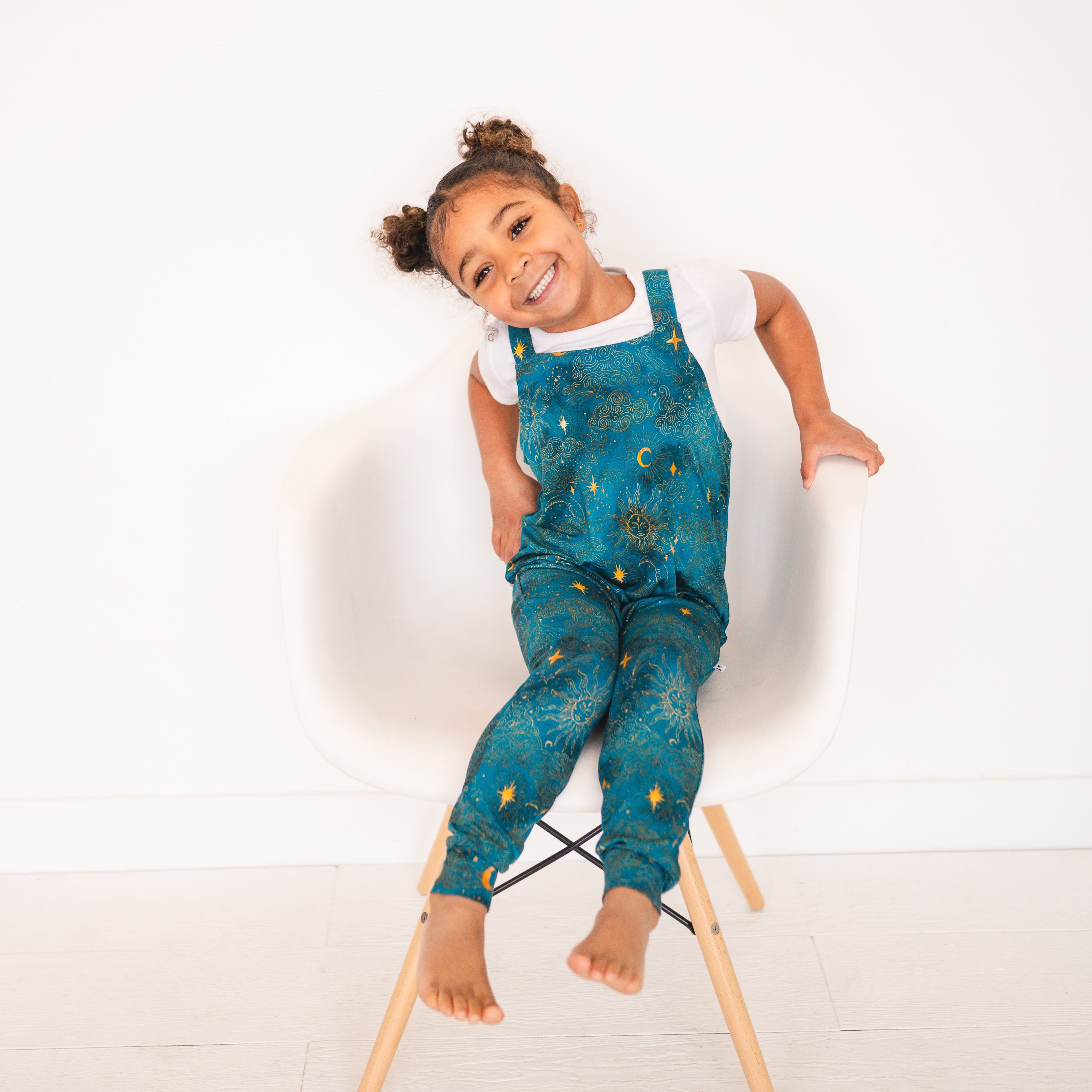 A young girl smiles while sitting in a chair wearing teal overalls with a celestial sun and moon pattern.