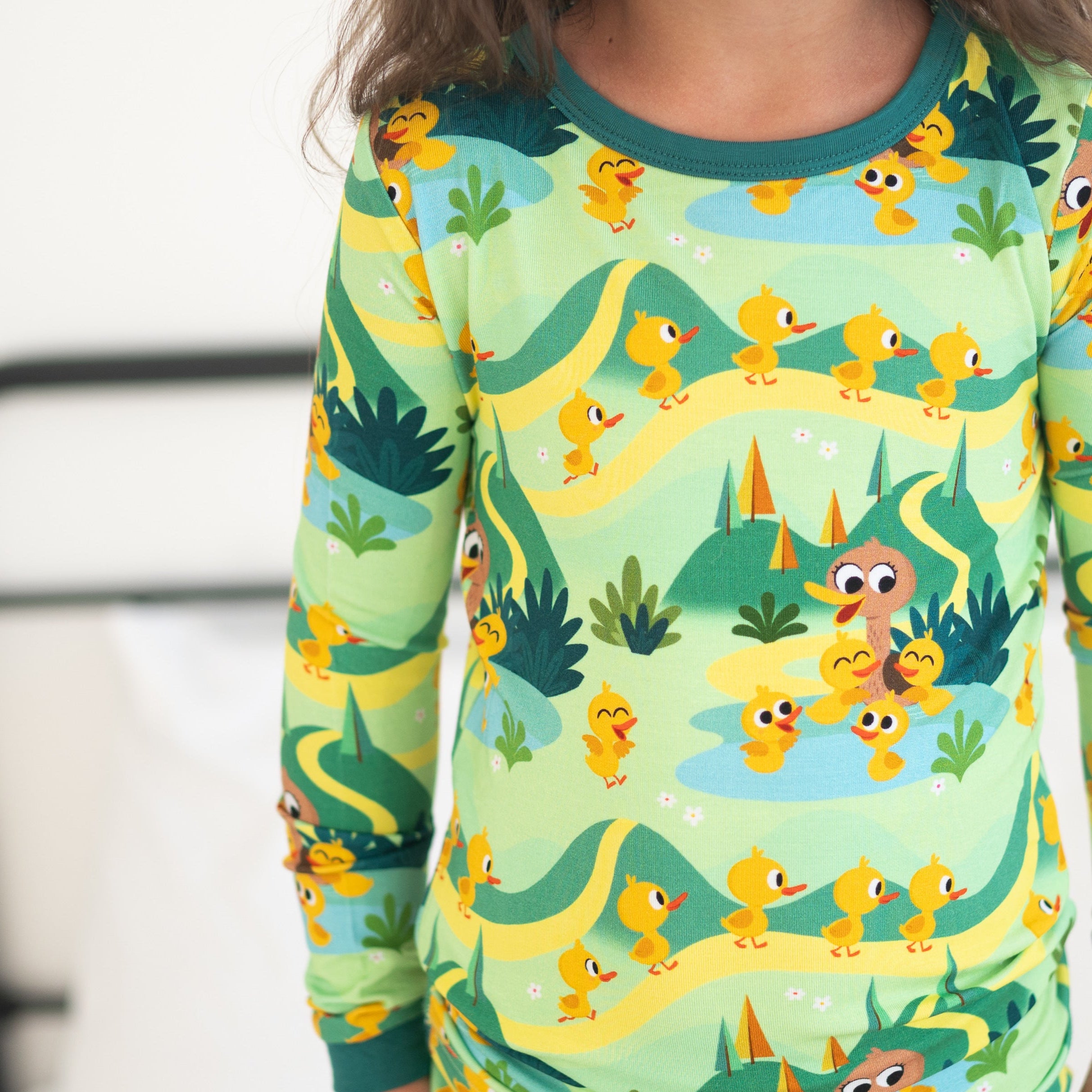 A child wears a light green pajama set with a pattern of yellow ducks and green hills.