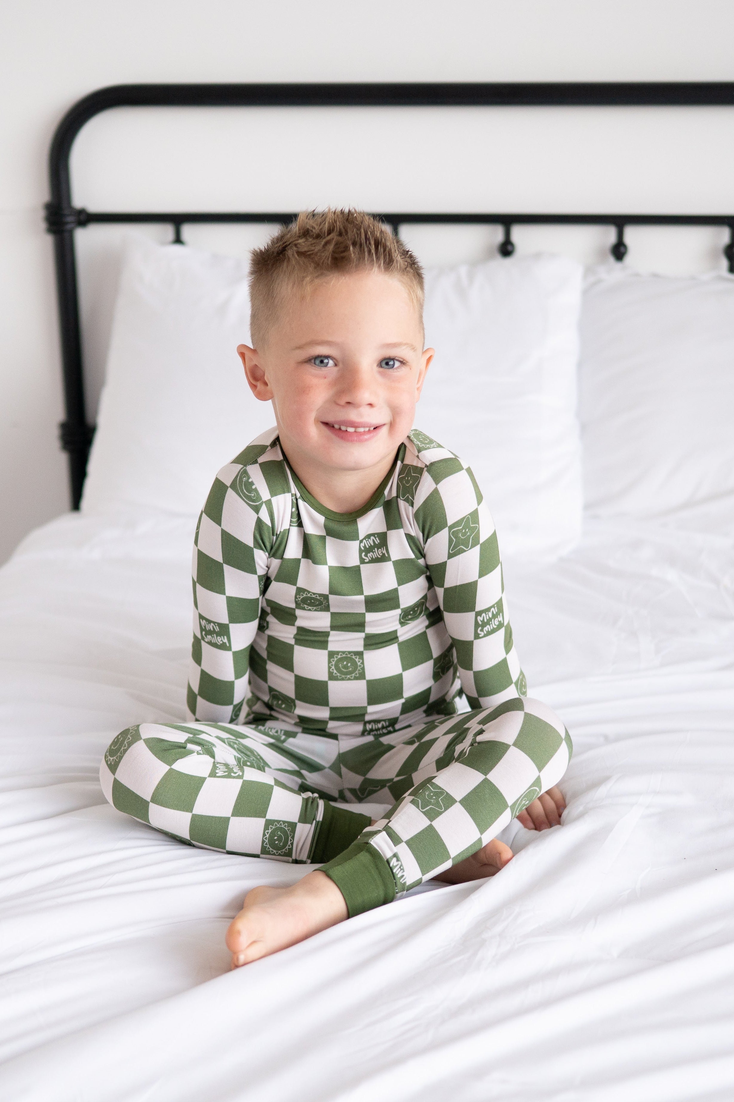 A young boy in green and white checkerboard pajamas with smiley faces sits on a bed.