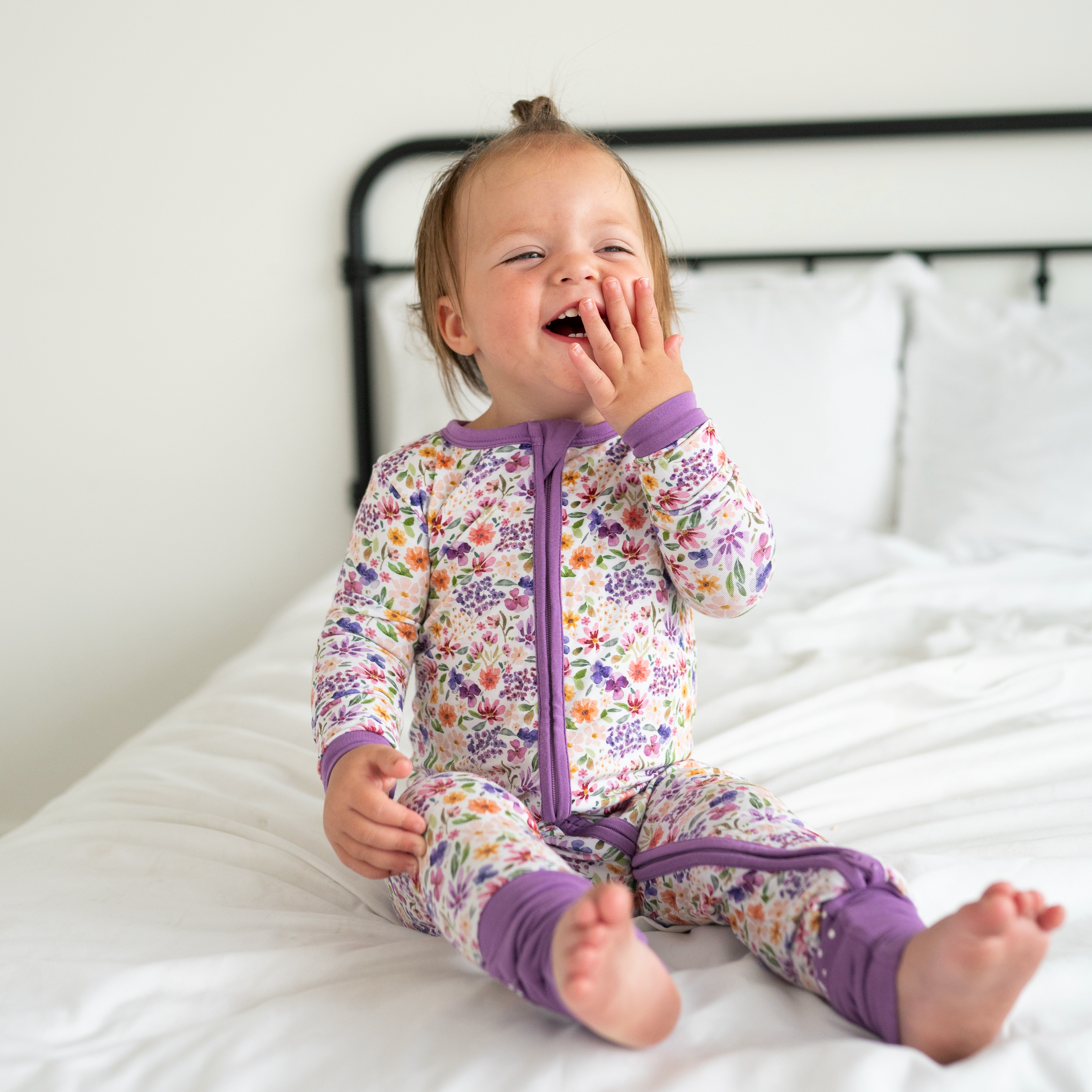A baby girl in a floral zip-up pajama set sits on a bed.