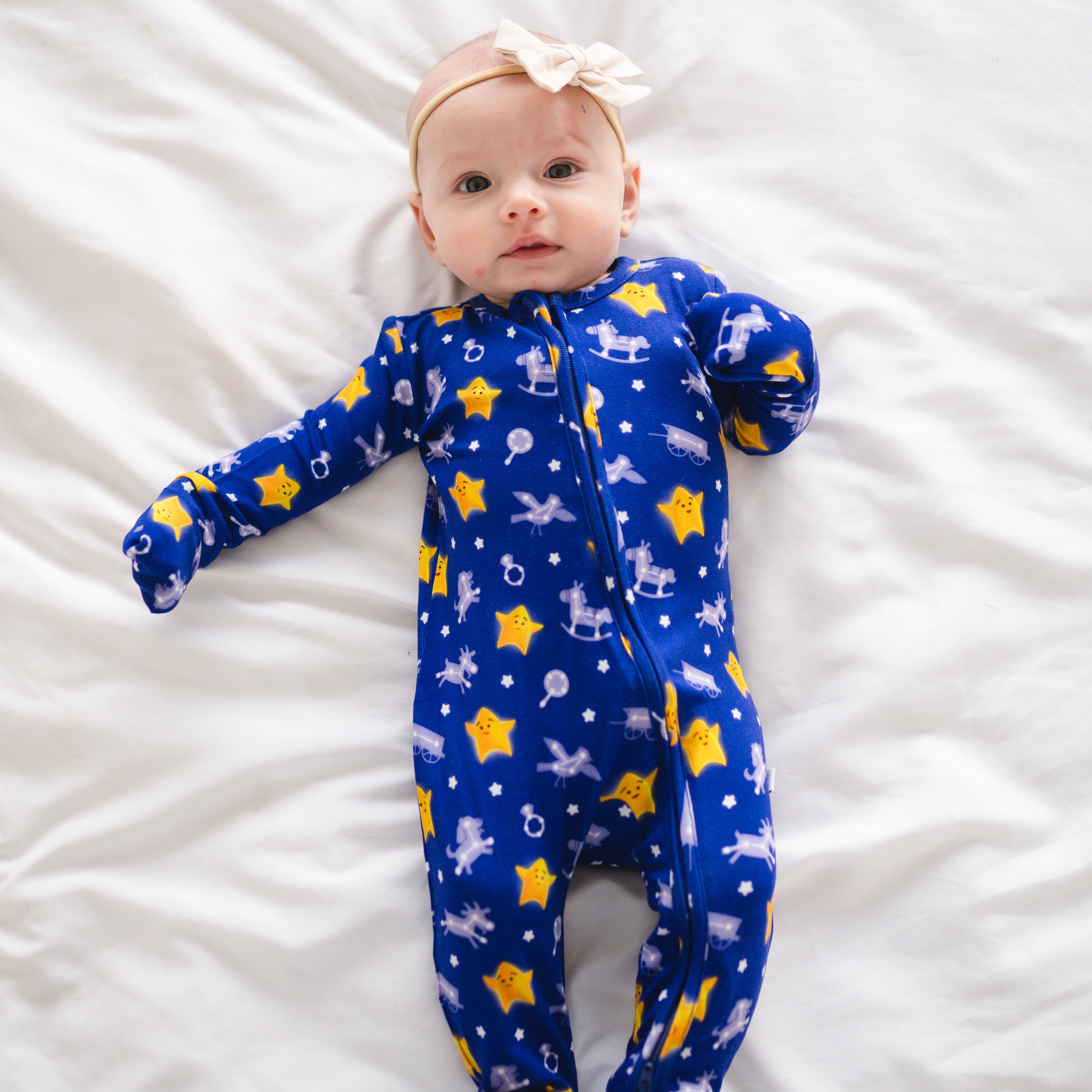 Baby in blue pajamas with stars and rocking horses, wearing a cream bow.