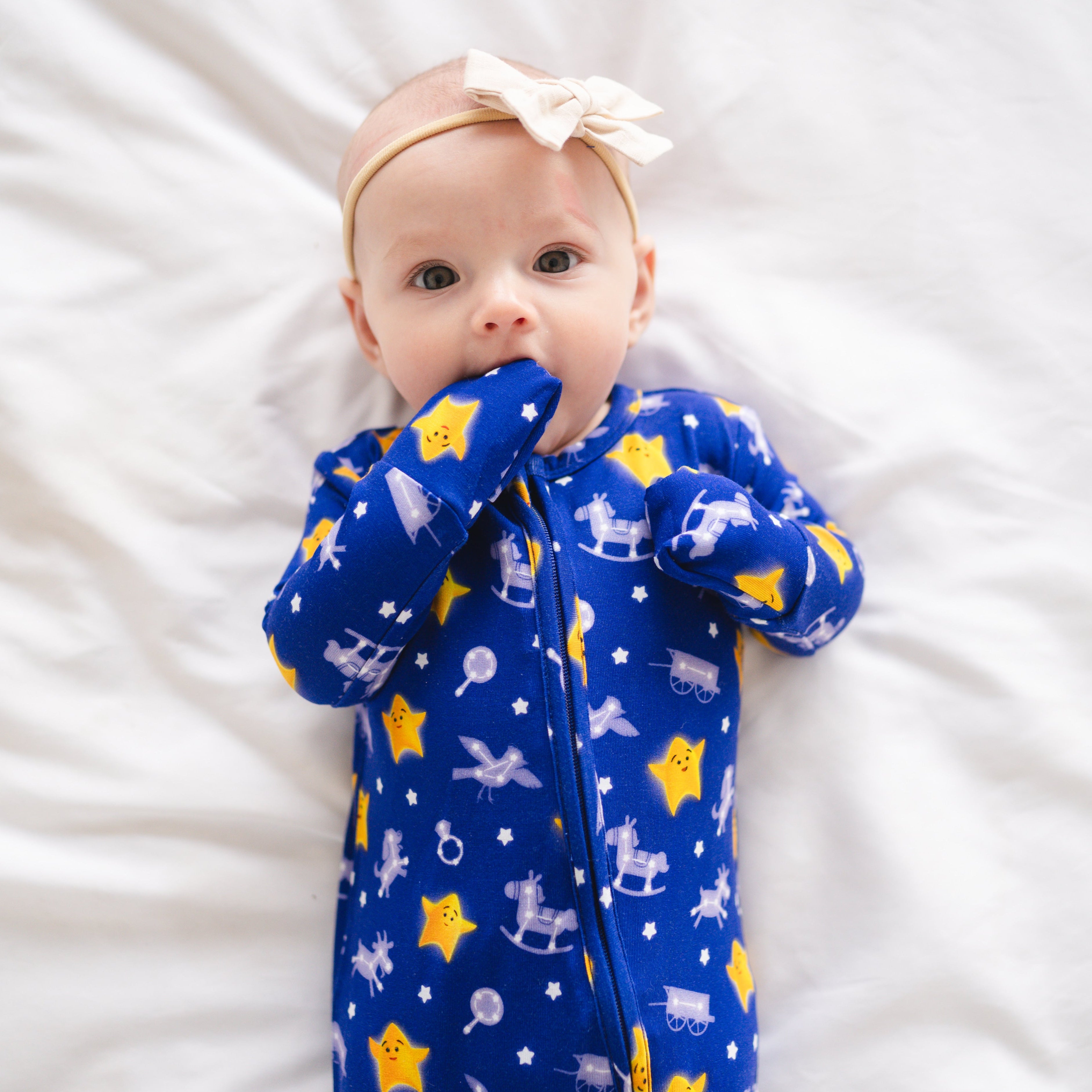 Baby in blue pajamas with stars and rocking horses, wearing a cream bow headband.