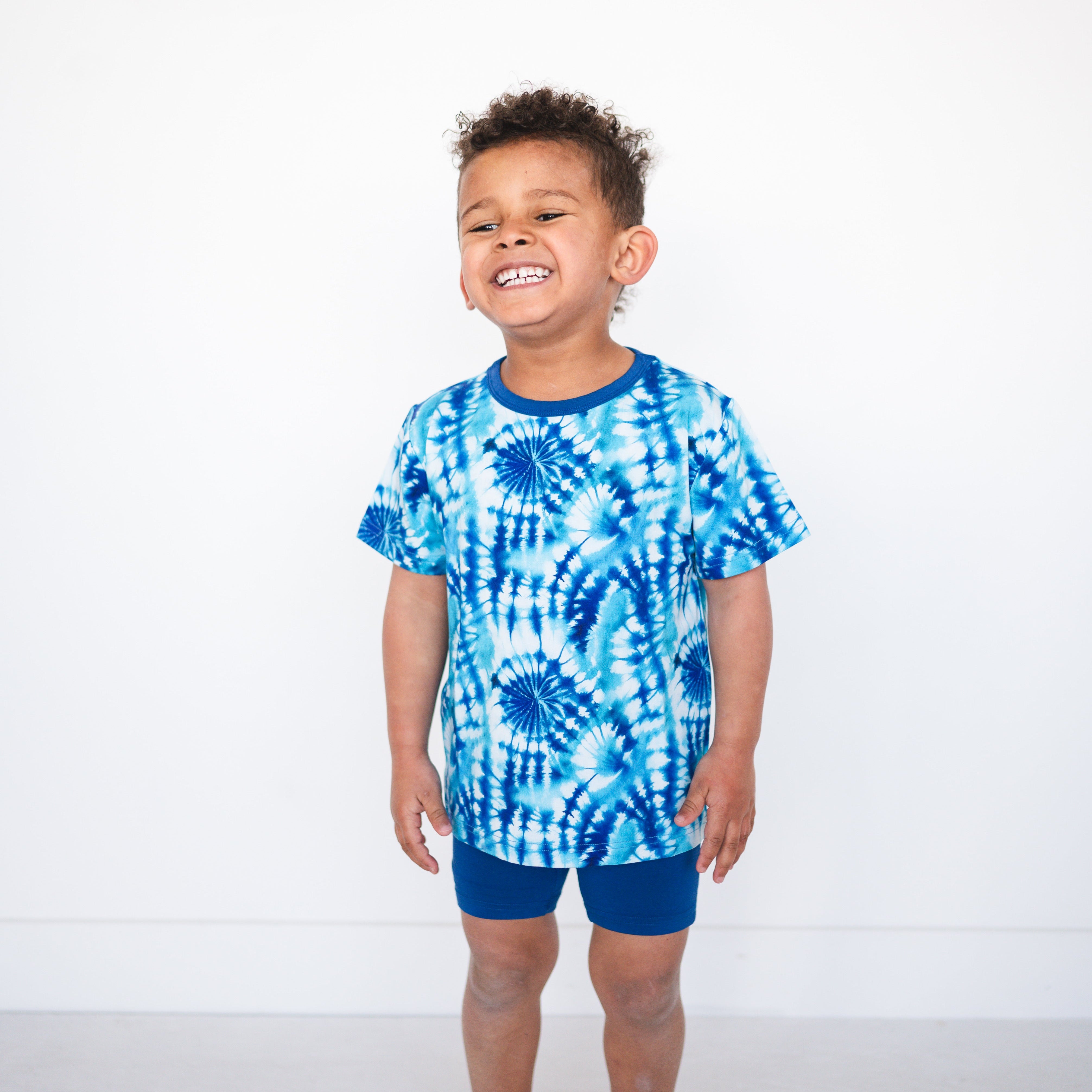 A young boy smiles while wearing a blue tie-dye t-shirt and blue shorts.