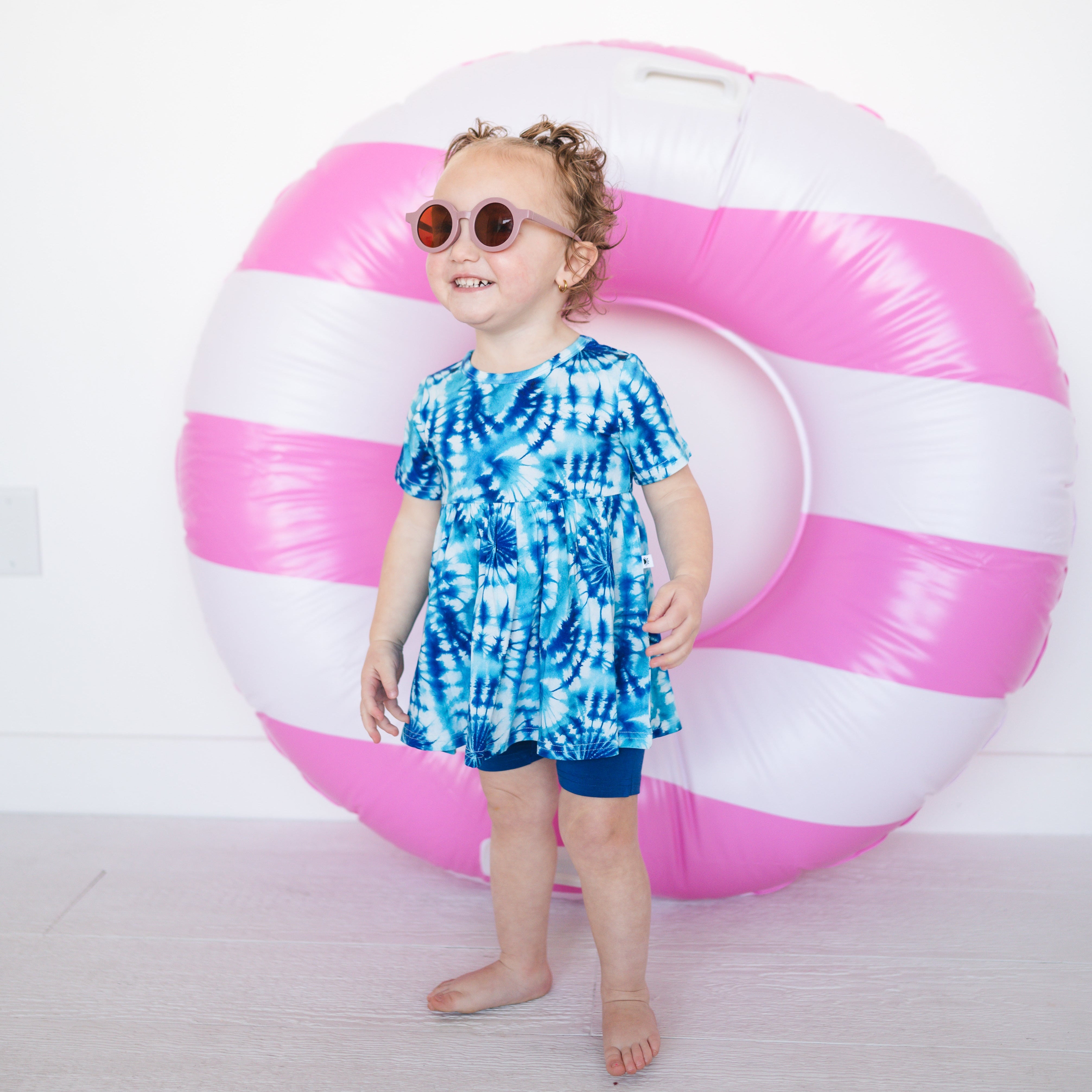 Toddler girl in a blue tie-dye peplum top and navy shorts, wearing sunglasses.