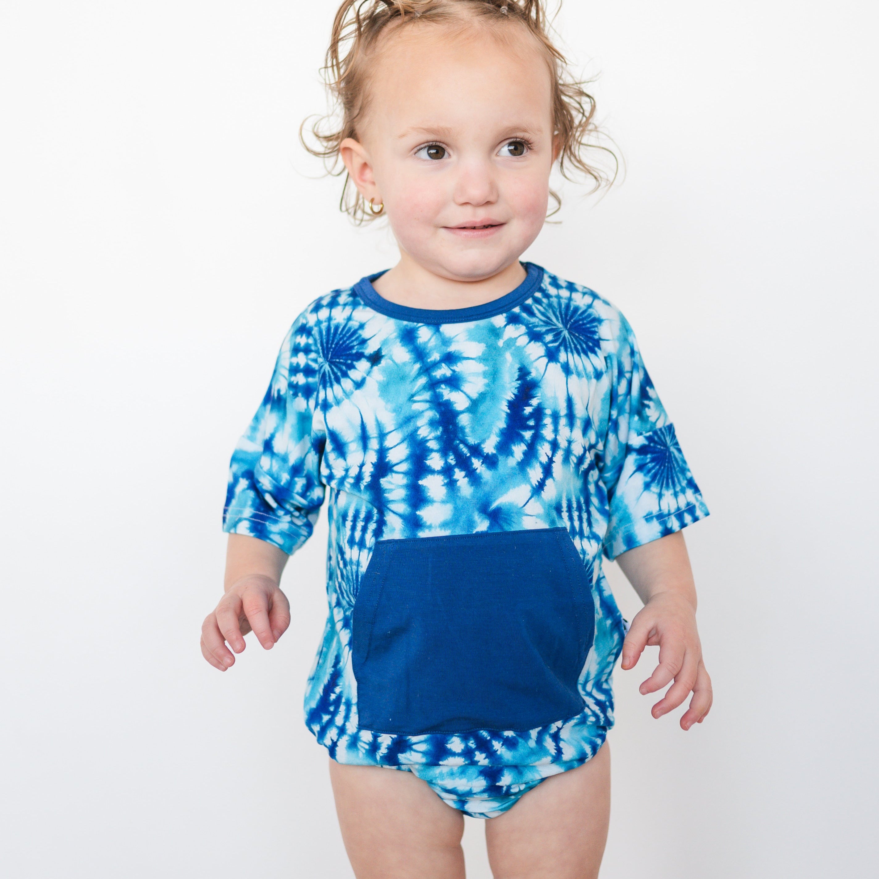 A toddler wears a blue tie-dye t-shirt romper with a solid blue pocket.