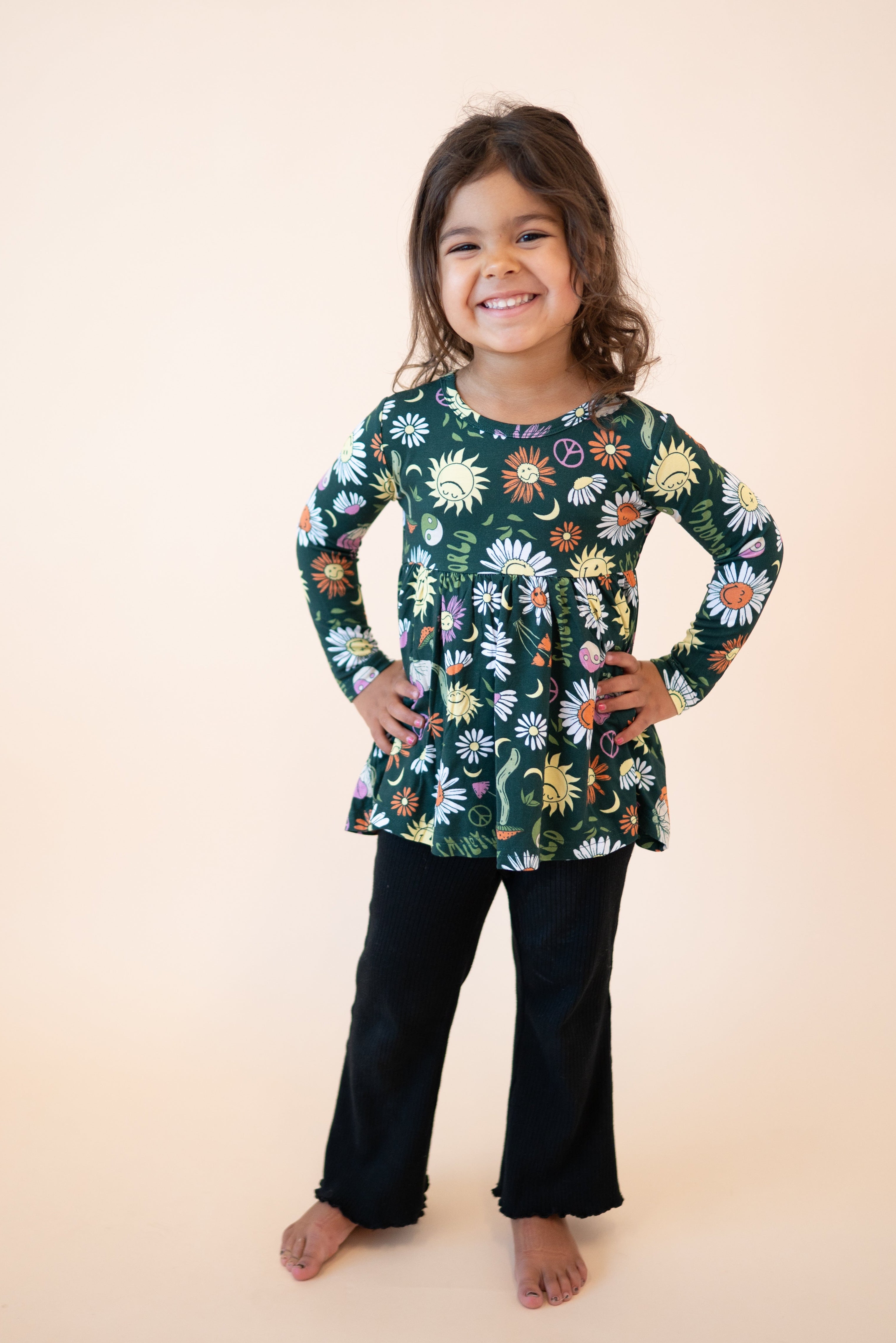 A young girl smiles wearing a long-sleeve green peplum top with a floral and smiley face print.