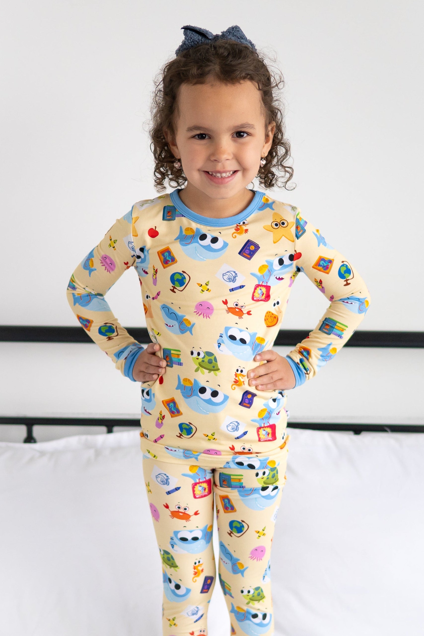 A young girl smiles in a yellow pajama set with a print of sharks and school supplies.