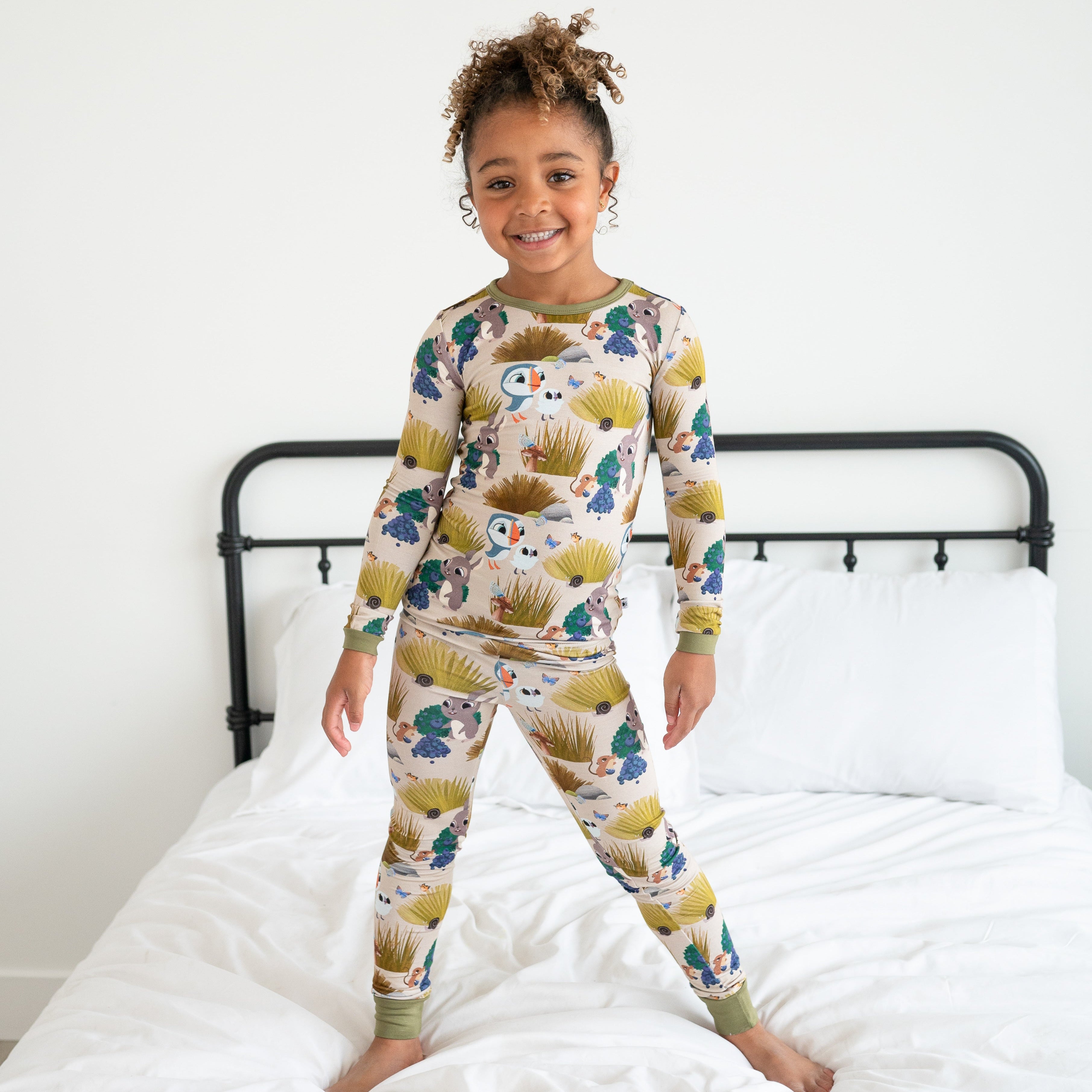 A young girl stands on a bed wearing a long-sleeved pajama set with a woodland animal print.