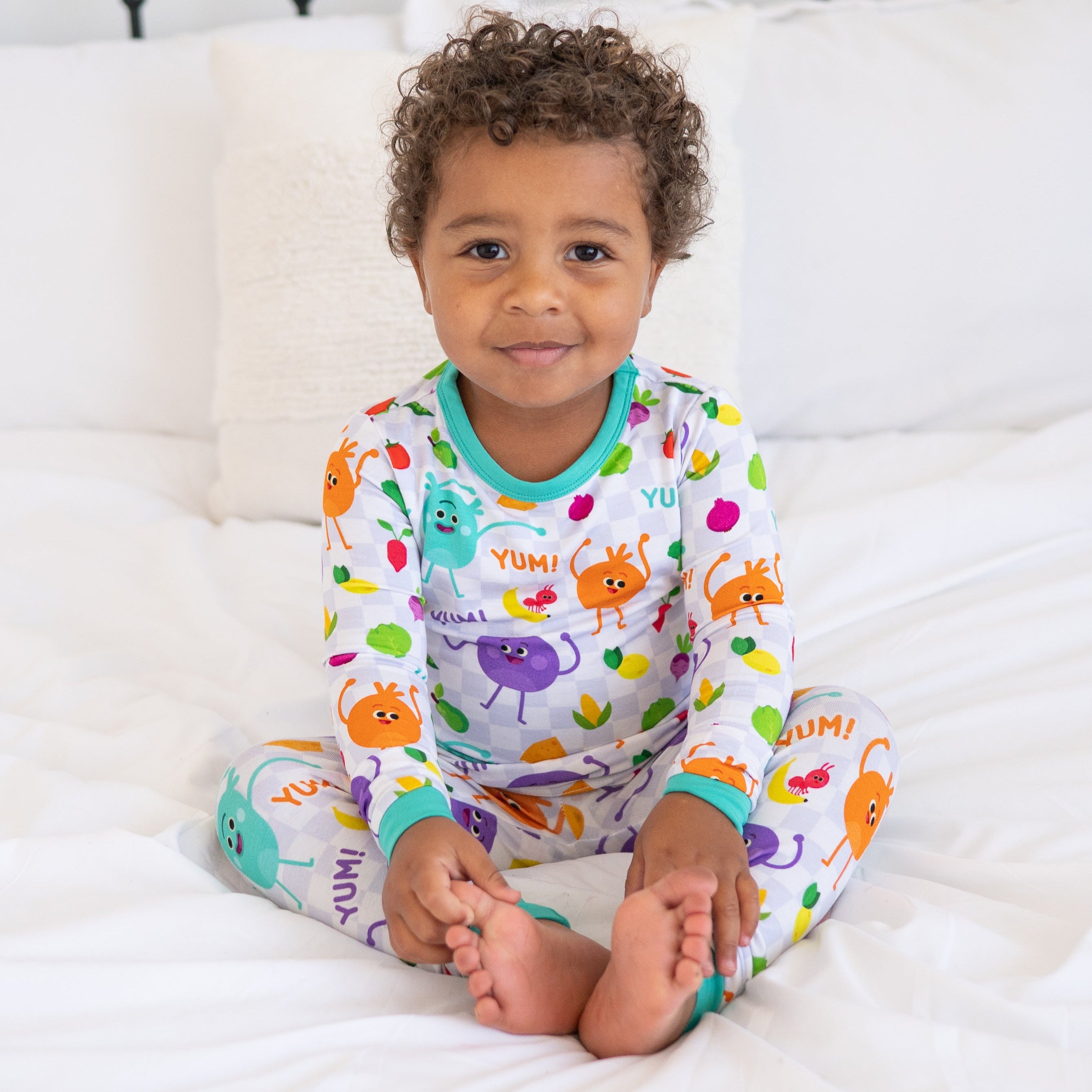 A young child sits on a bed wearing pajamas with a checkerboard pattern and cartoon vegetables.