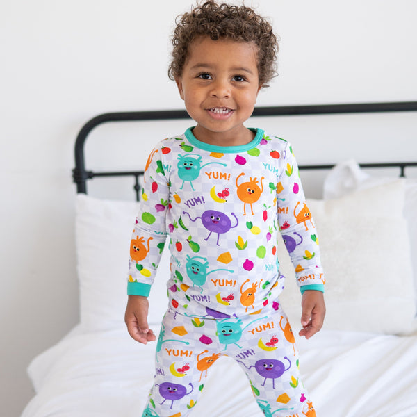 A young boy smiles while wearing white pajamas with a checkerboard pattern and colorful fruit and vegetable characters.