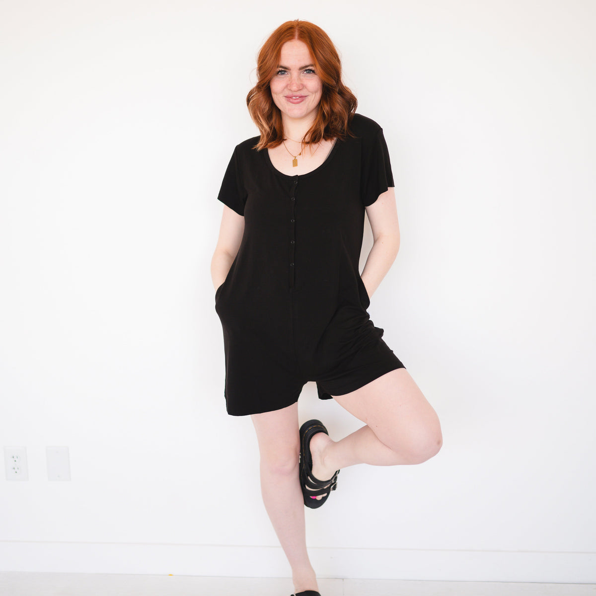 A woman in a black short-sleeve romper with a gold necklace stands against a white wall.