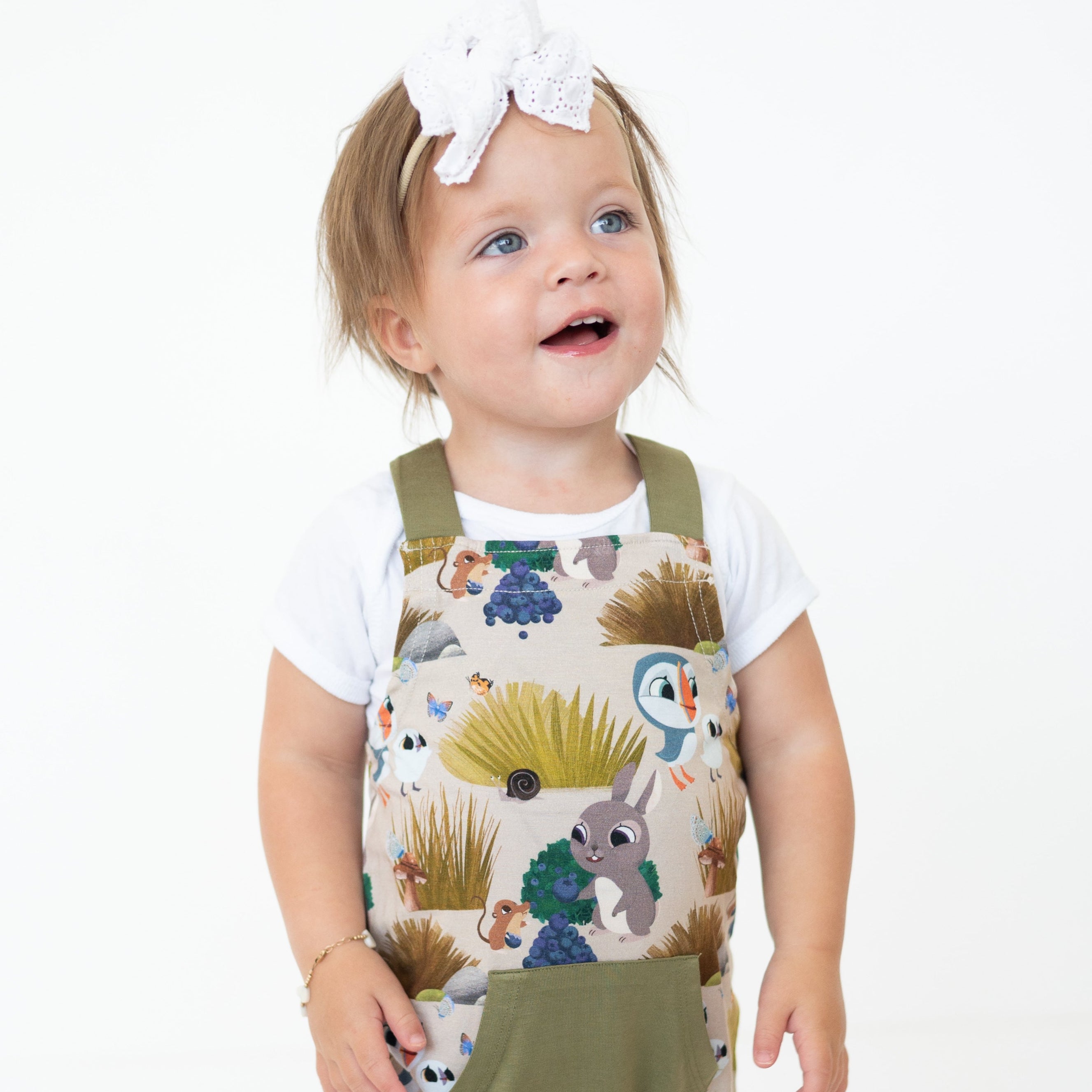 A toddler girl wears Puffin Rock™ bamboo overalls with a white shirt and a white bow.