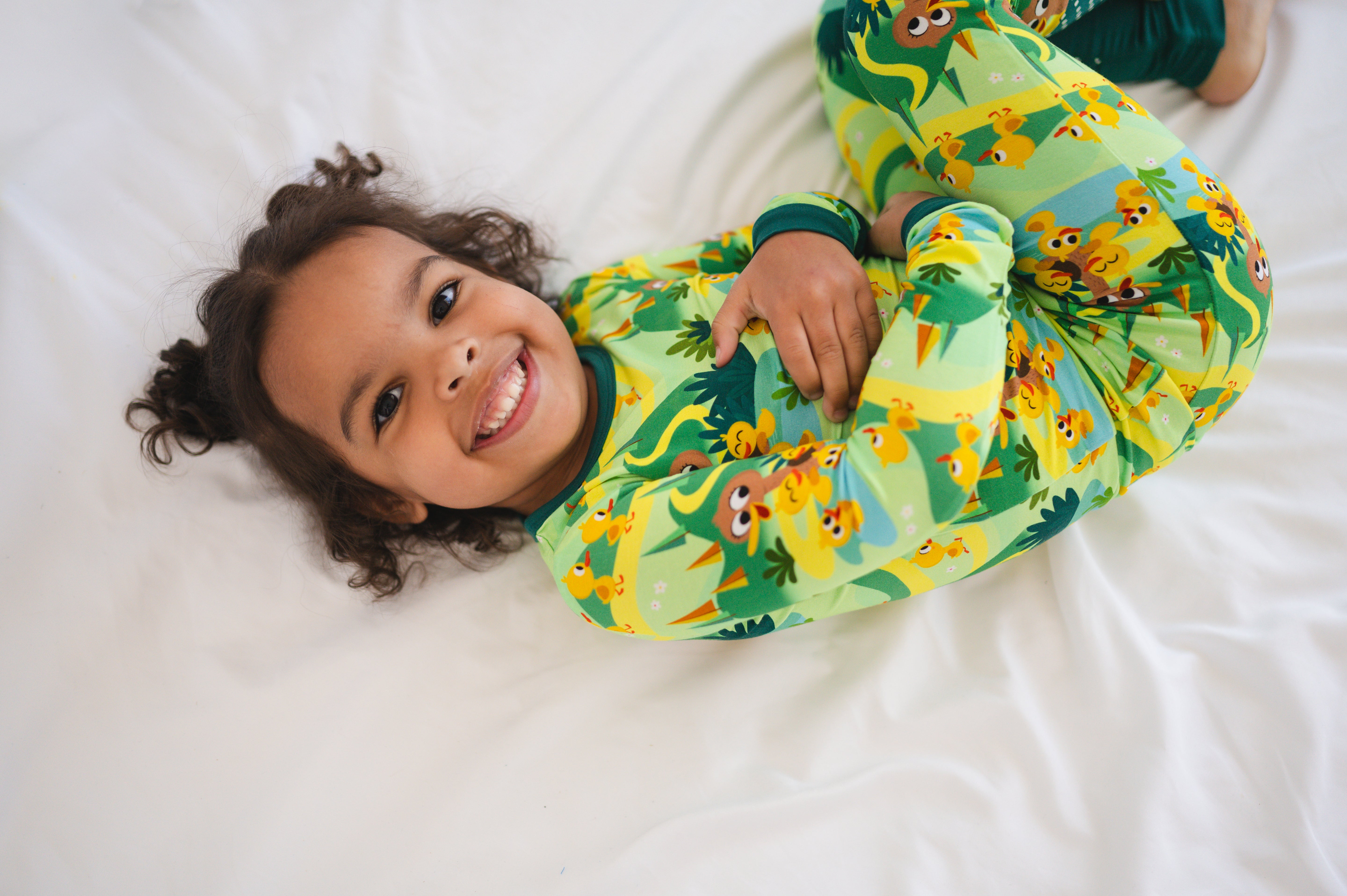 A child in green pajamas with a duck print smiles while lying on a white sheet.
