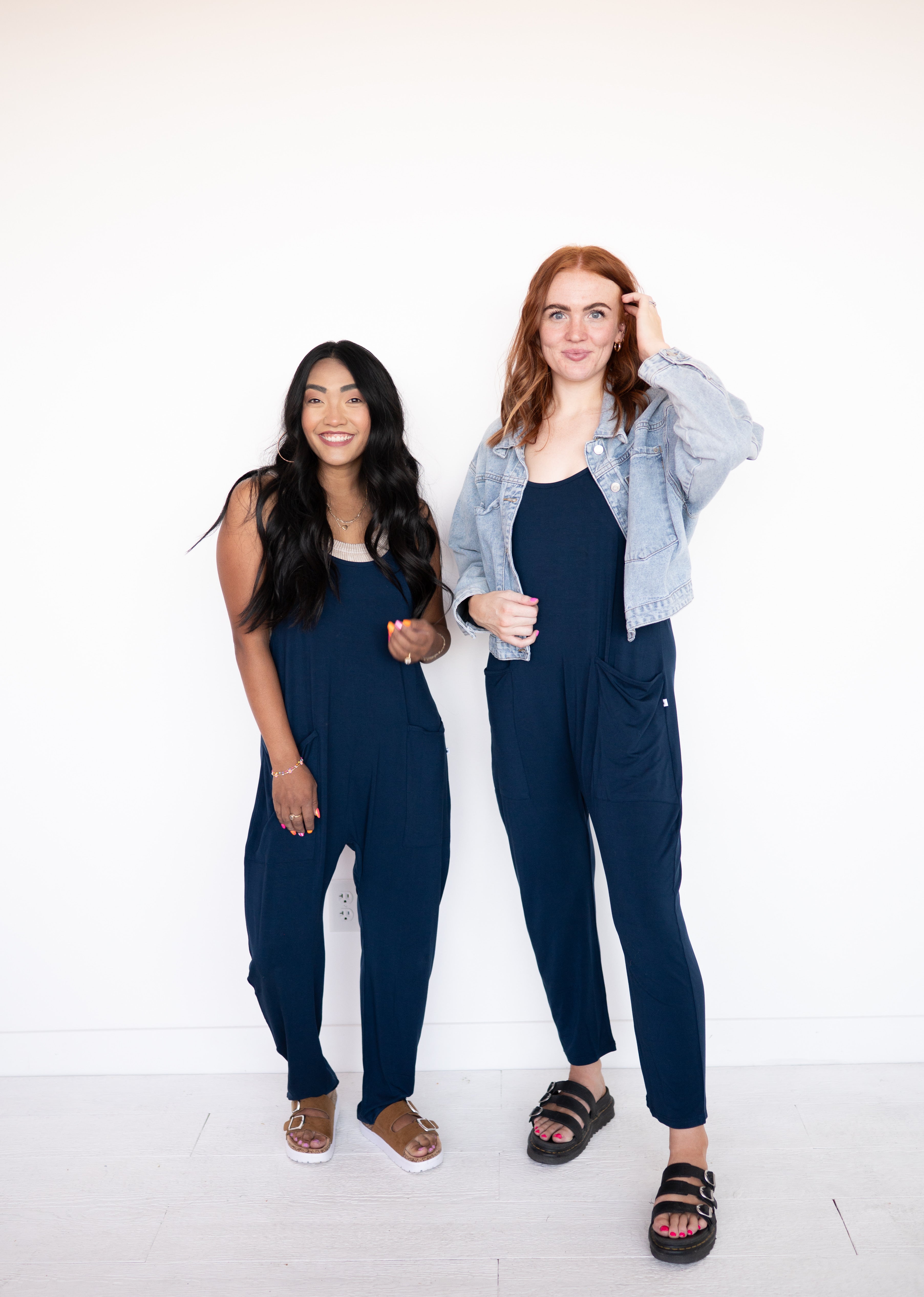 Two women in navy bamboo overalls and one in a denim jacket stand against a white wall.