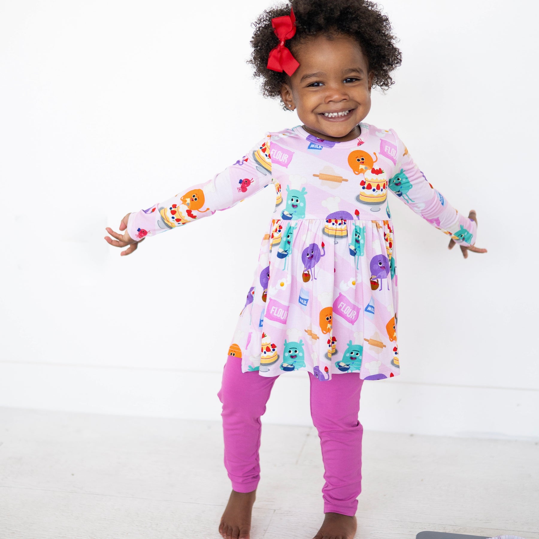 A young girl smiles wearing a pink long-sleeve peplum top with a baking-themed print and purple leggings.