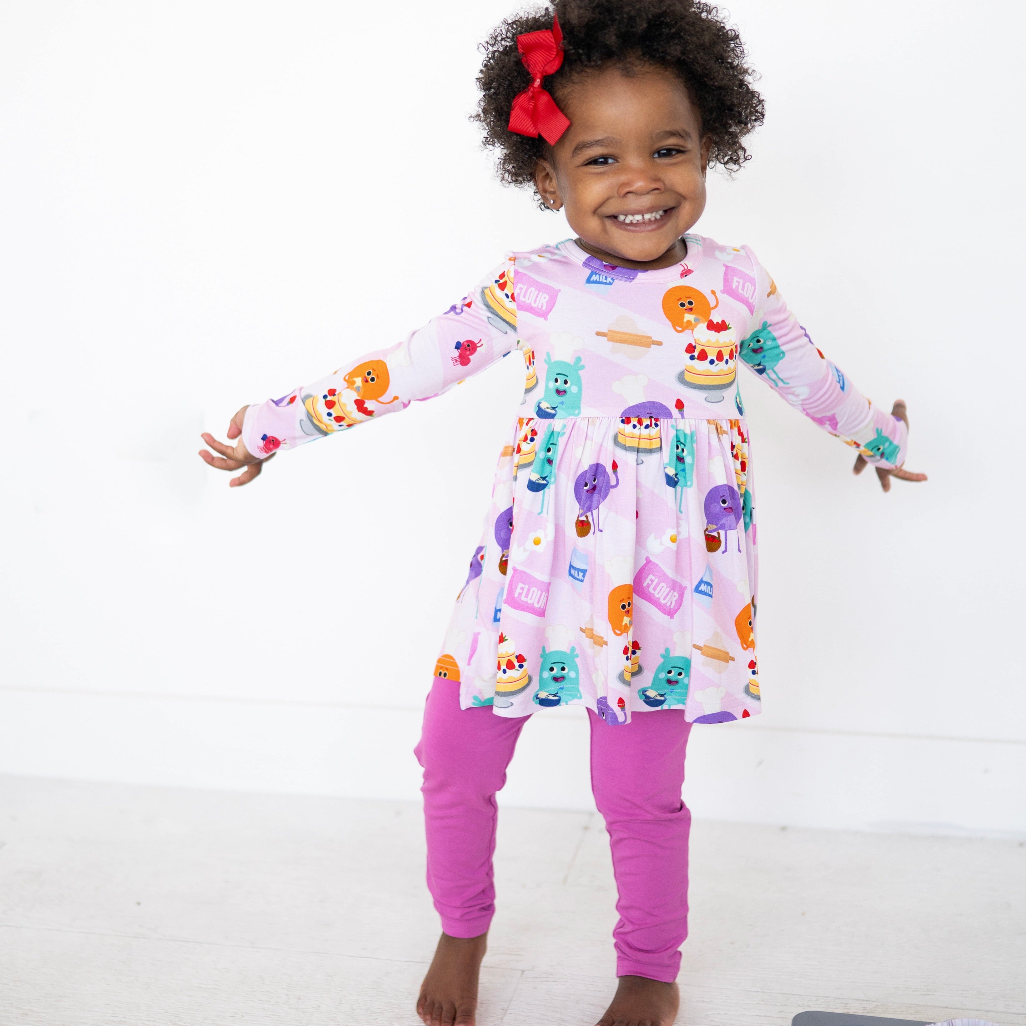 A young girl smiles wearing a pink long-sleeve peplum top with a baking-themed print and purple leggings.