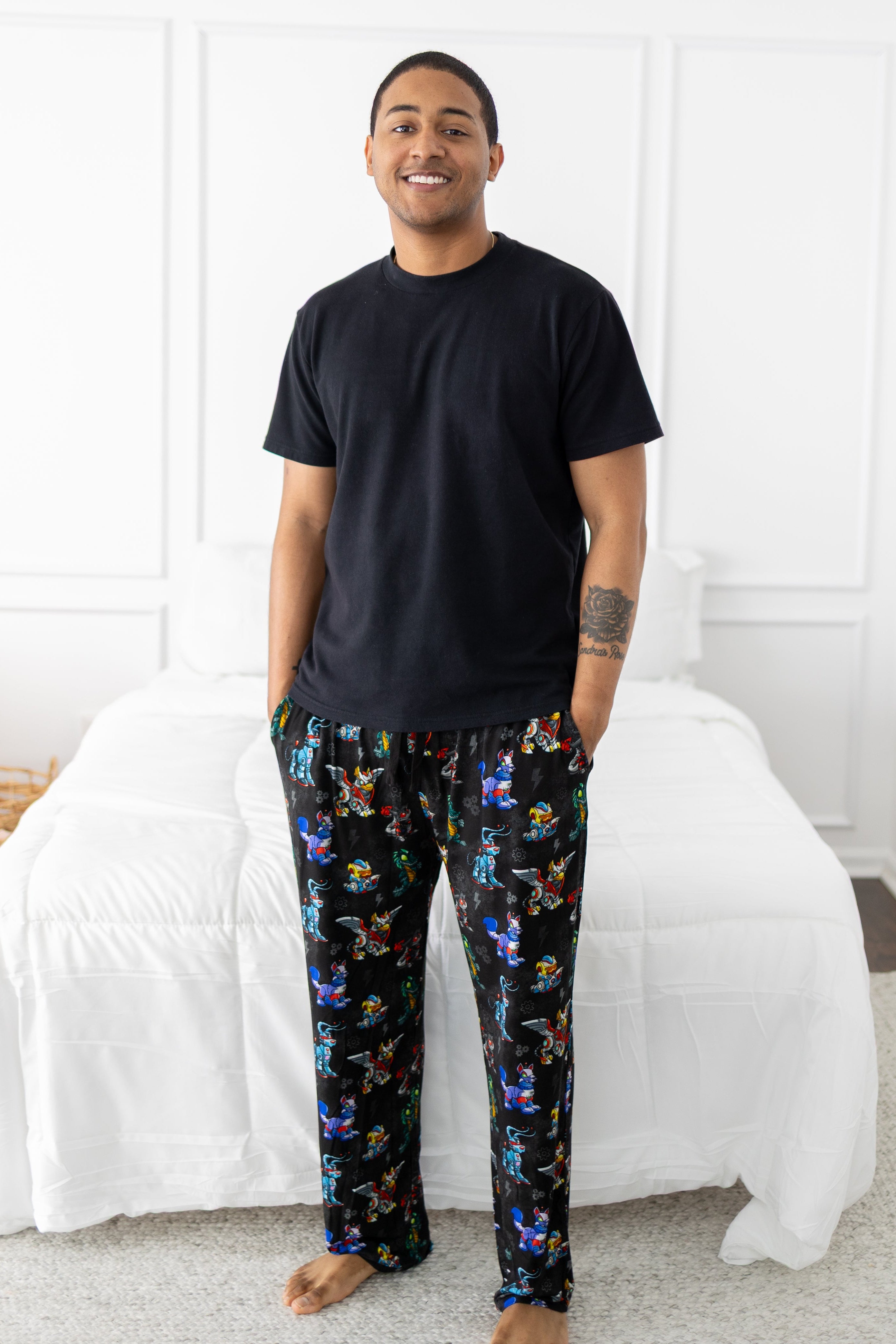 A man stands in a bedroom wearing black Neopets Robot print sleep pants and a black t-shirt.