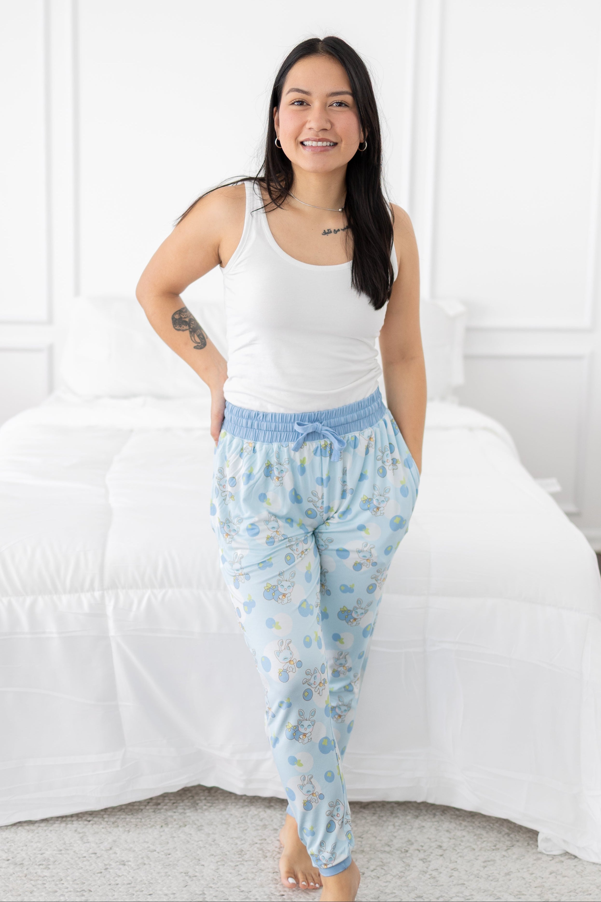 A woman wears light blue Neopets blueberry baby patterned joggers with a white tank top.