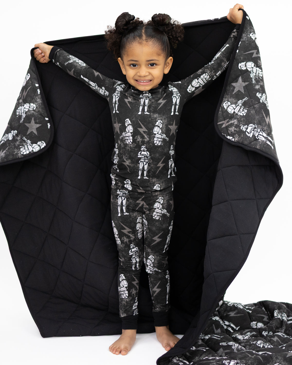 A child in Stormtrooper pajamas holds a black quilted blanket with a Stormtrooper print.