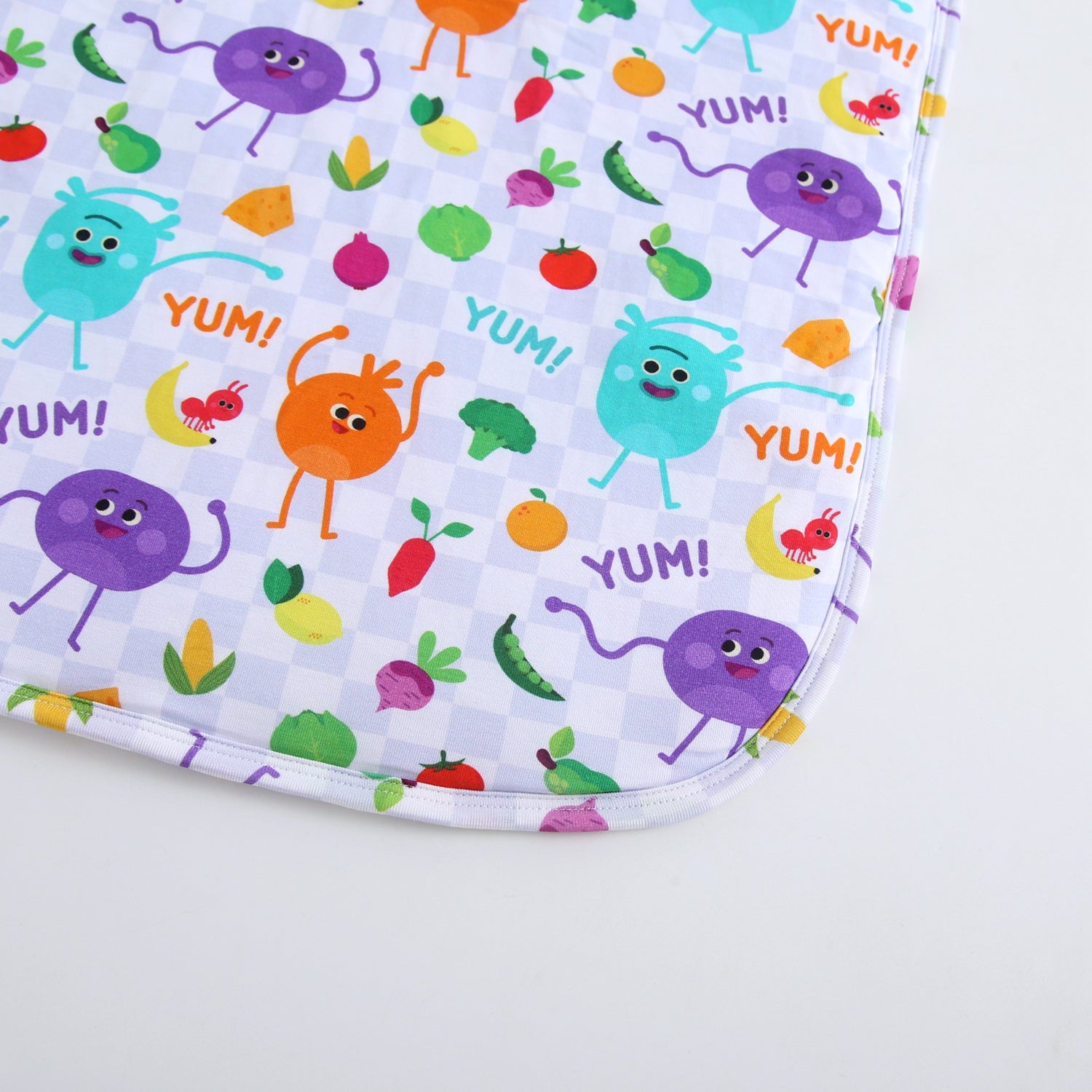 A white blanket with a checkered pattern featuring cartoon vegetables and the word "YUM".