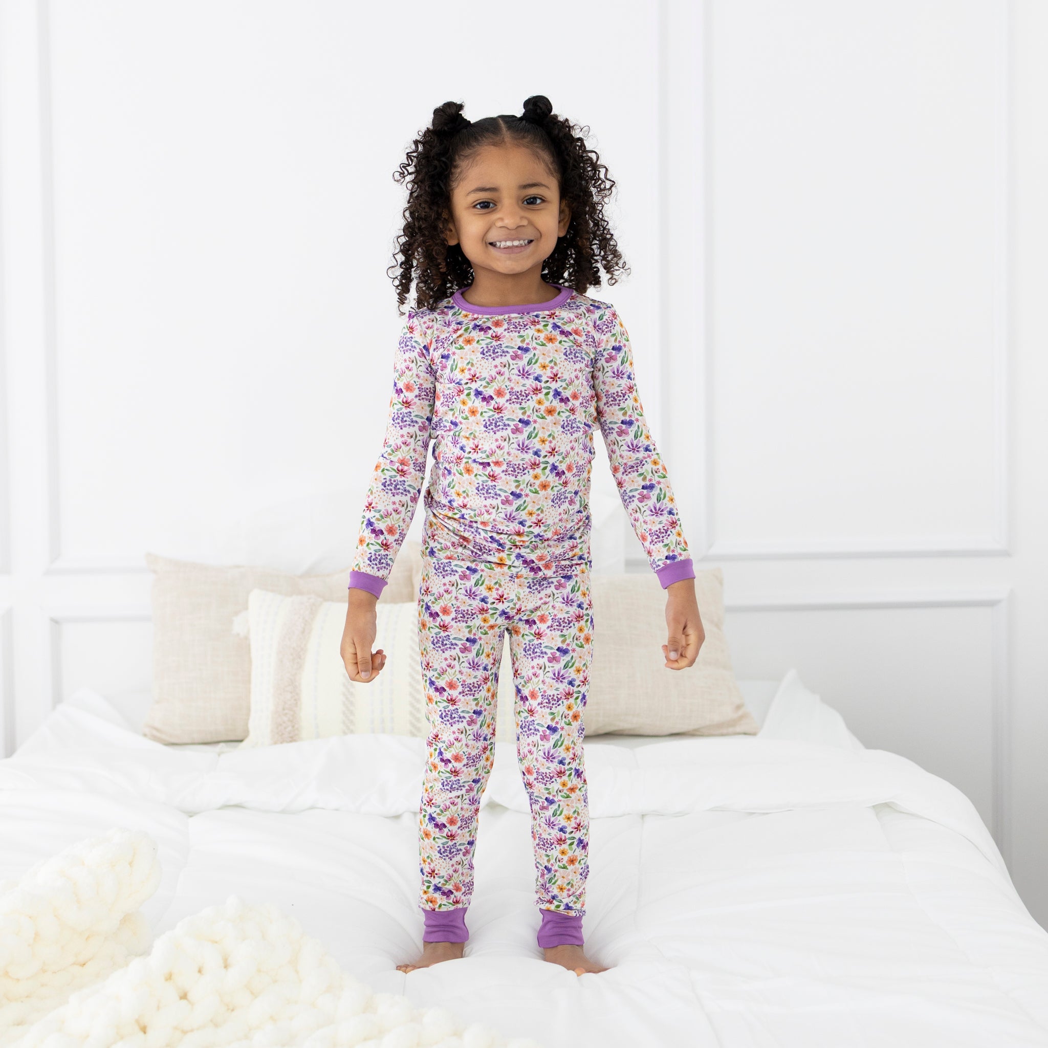 A child stands on a bed wearing a floral print long-sleeve pajama set with purple trim.