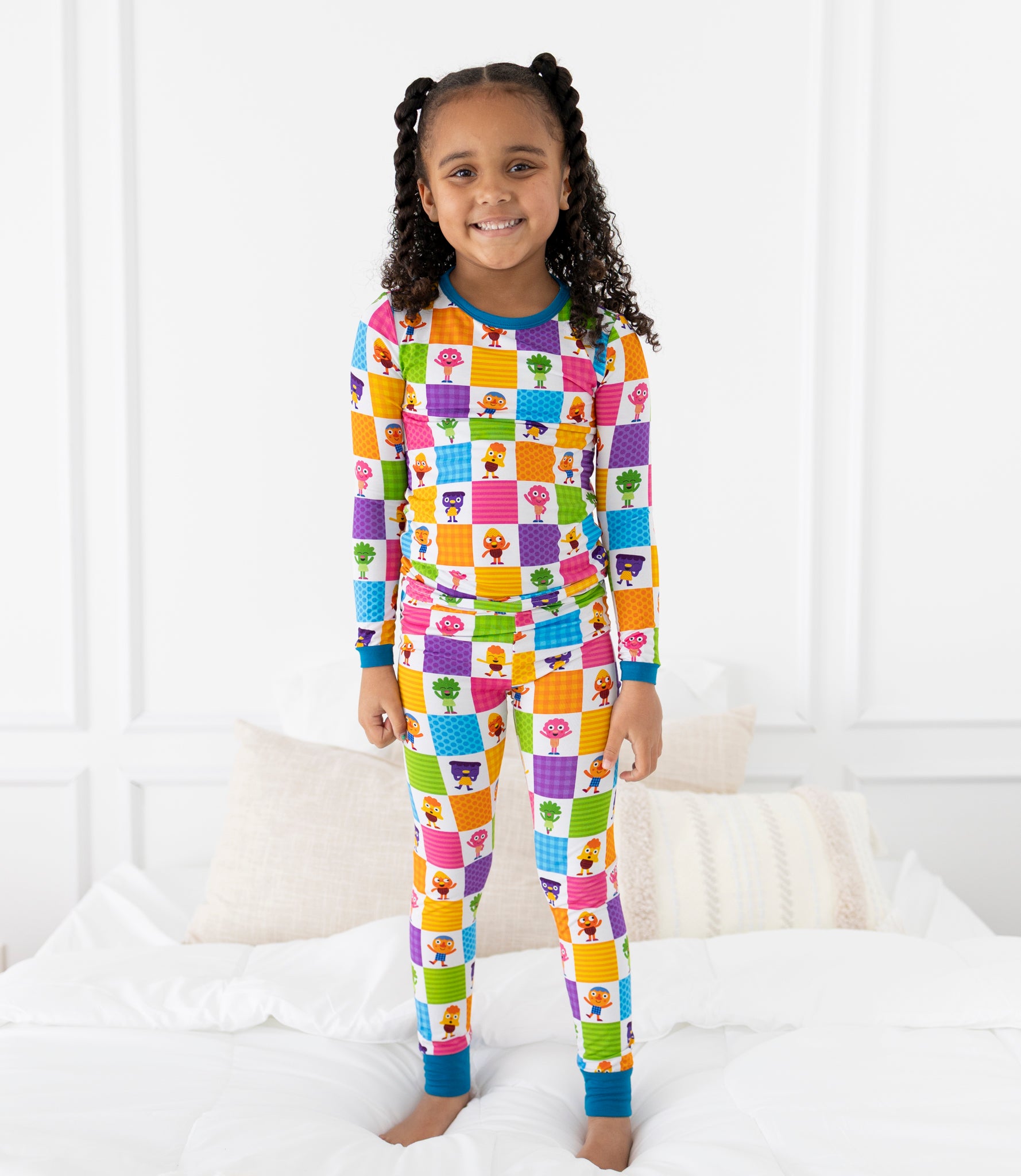 A young girl stands on a bed wearing colorful pajamas with a checkered pattern of cartoon characters.