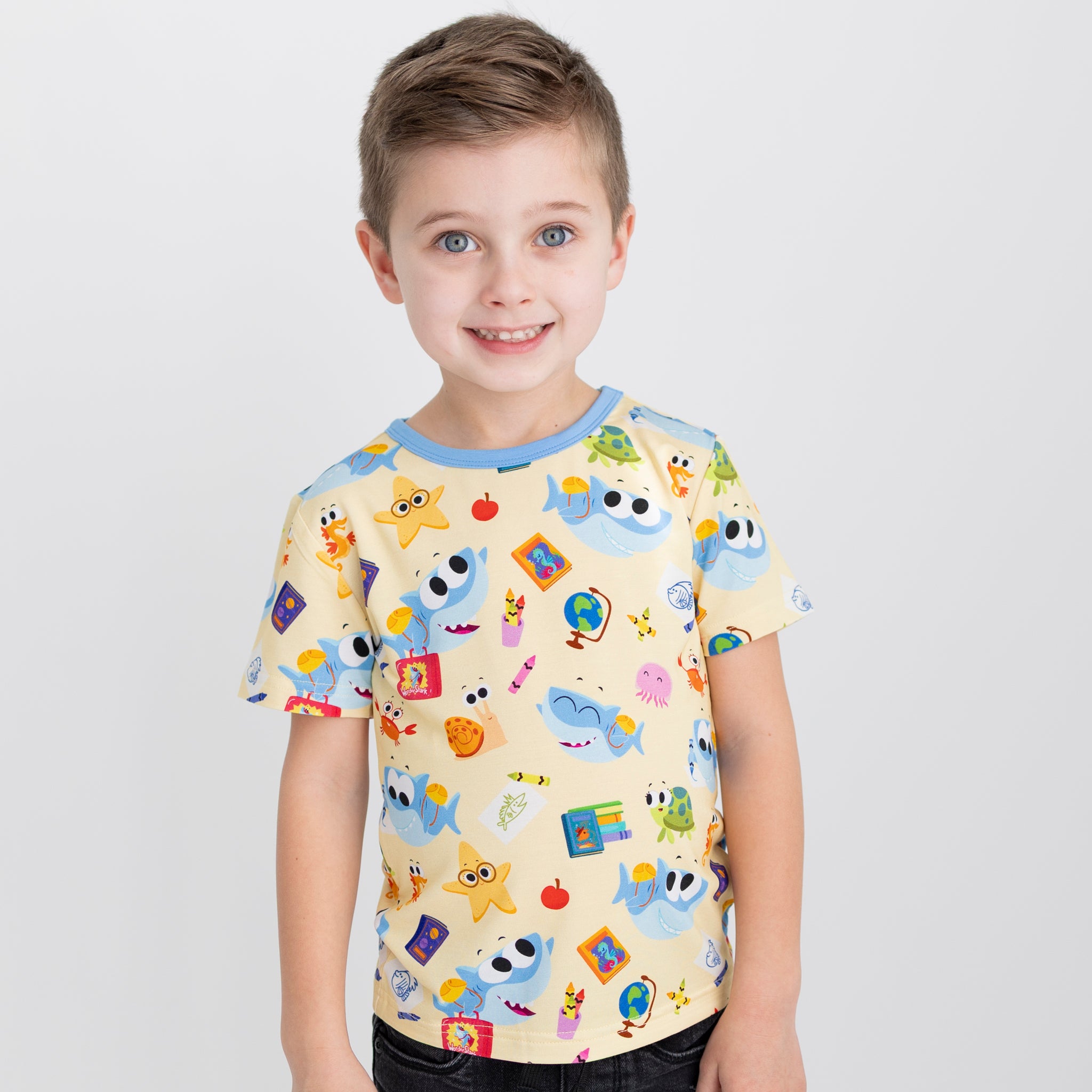 A young boy smiles wearing a yellow T-shirt featuring a Super Simple Finny the Shark school print.