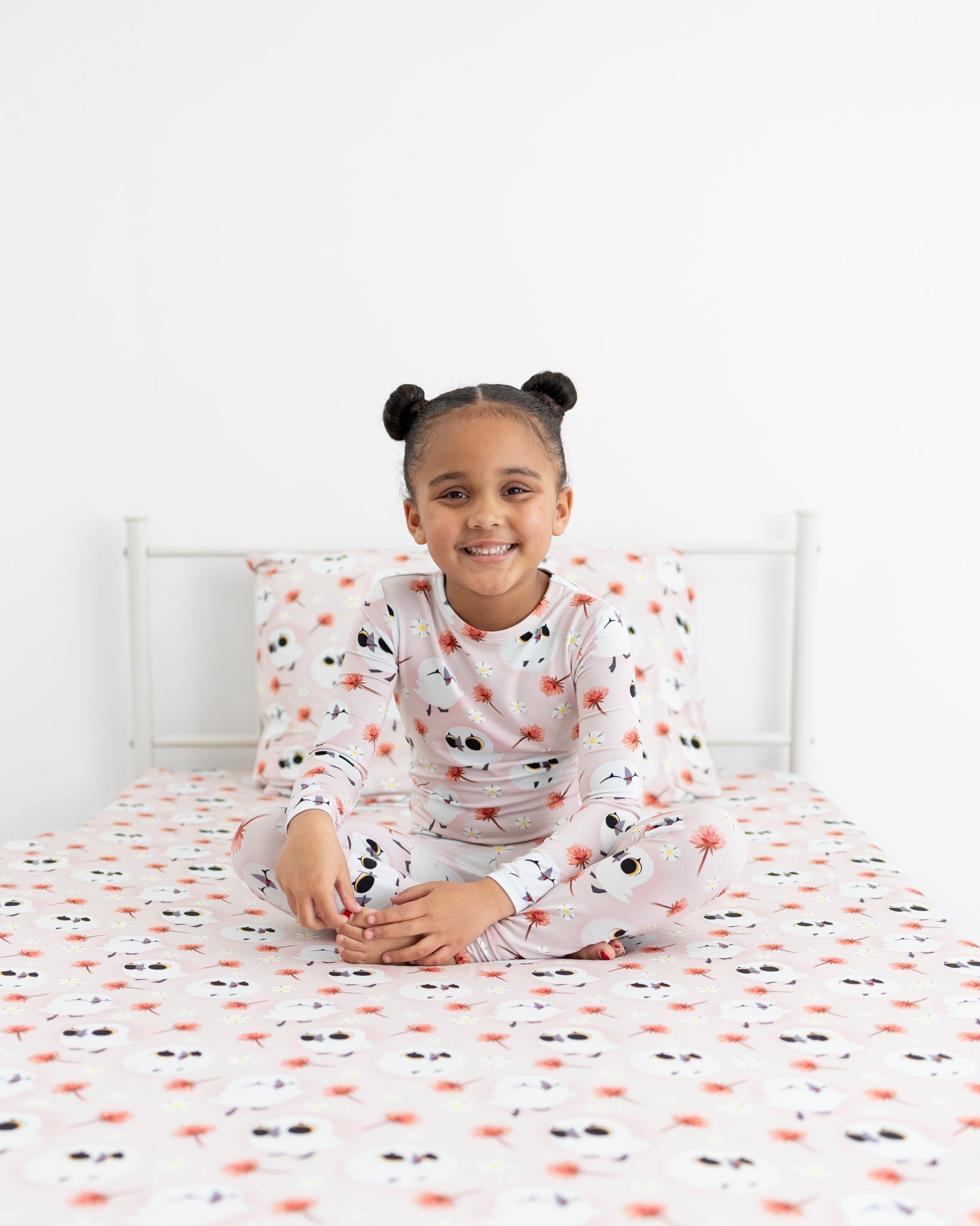 A young girl sits cross-legged on a bed wearing a pink pajama set with a puffin print.