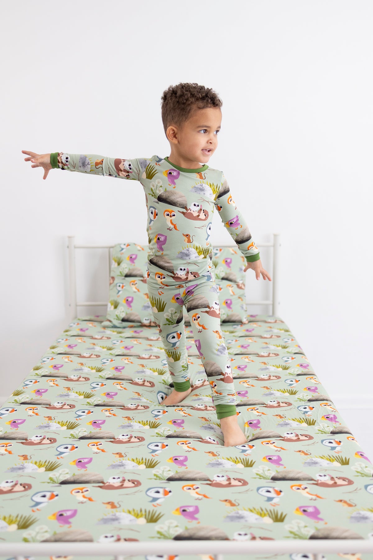 A young boy wears a light green pajama set with a Puffin Rock™ print, standing on a matching bed.