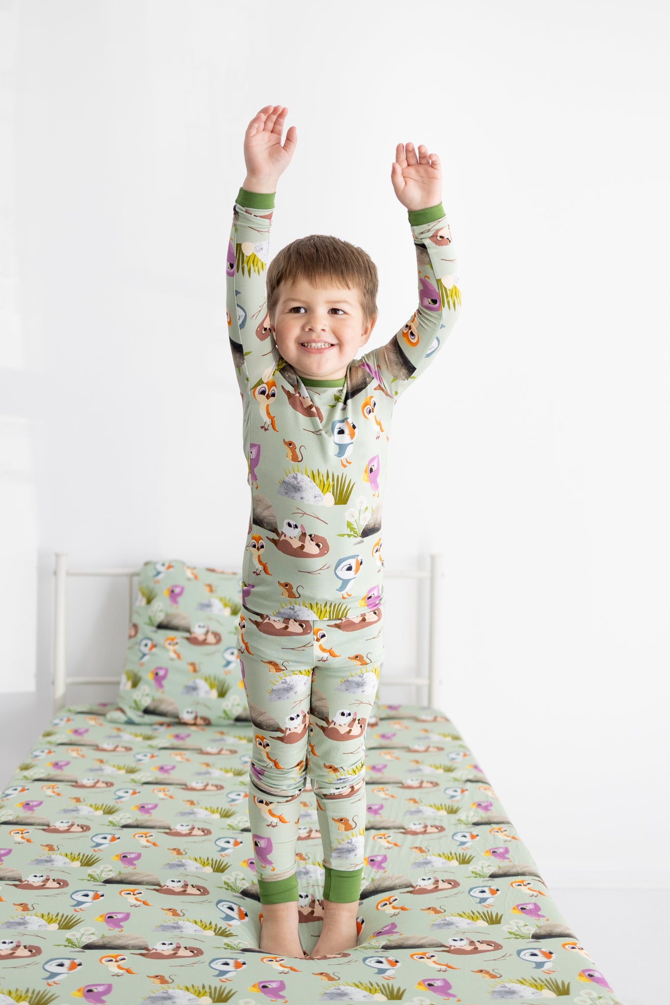 A young boy in Puffin Rock pajamas stands on a bed with matching bedding.