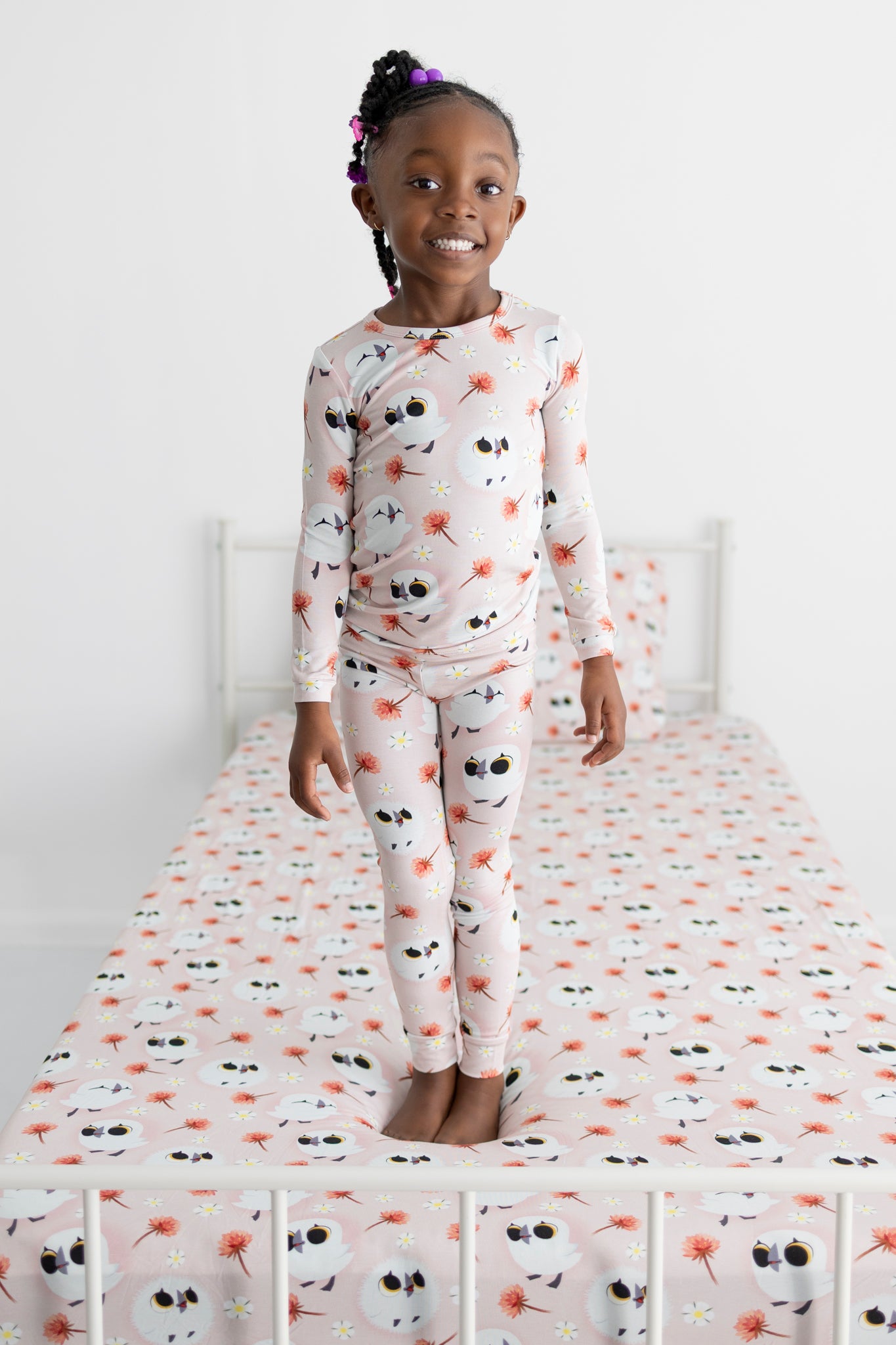 A young girl stands on a bed wearing pink pajamas with a puffin print.