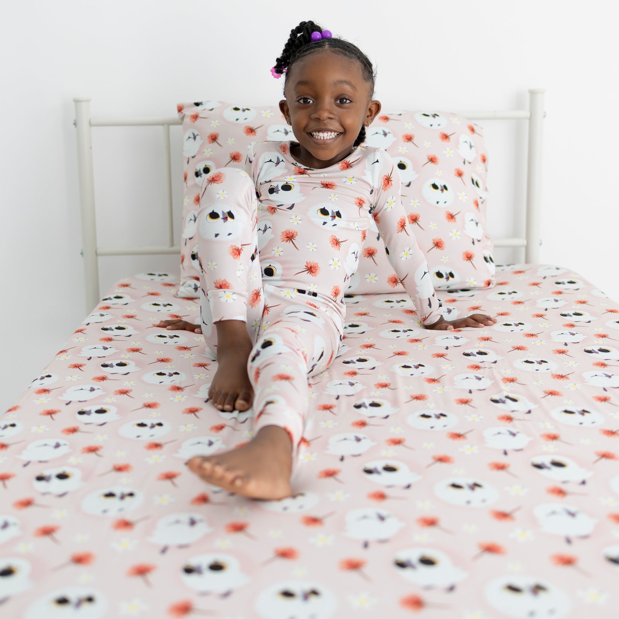 A young girl in pink owl-print pajamas sits on a bed with matching bedding.