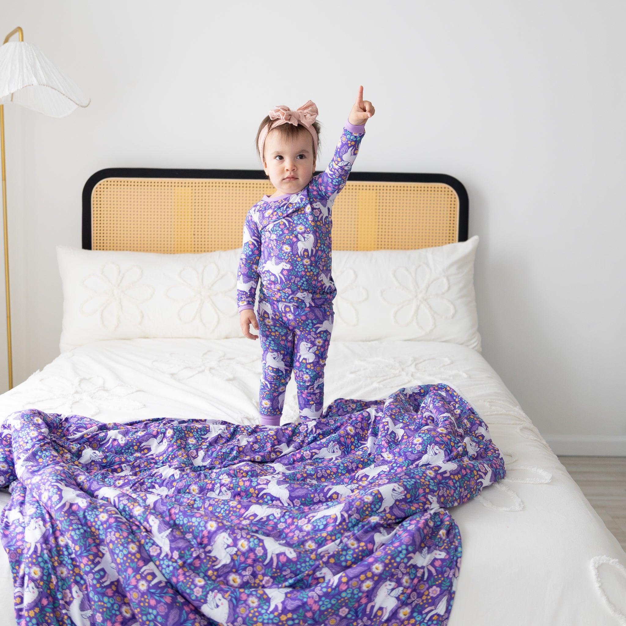 A toddler stands on a bed wearing matching purple unicorn-print pajamas and a bow headband.