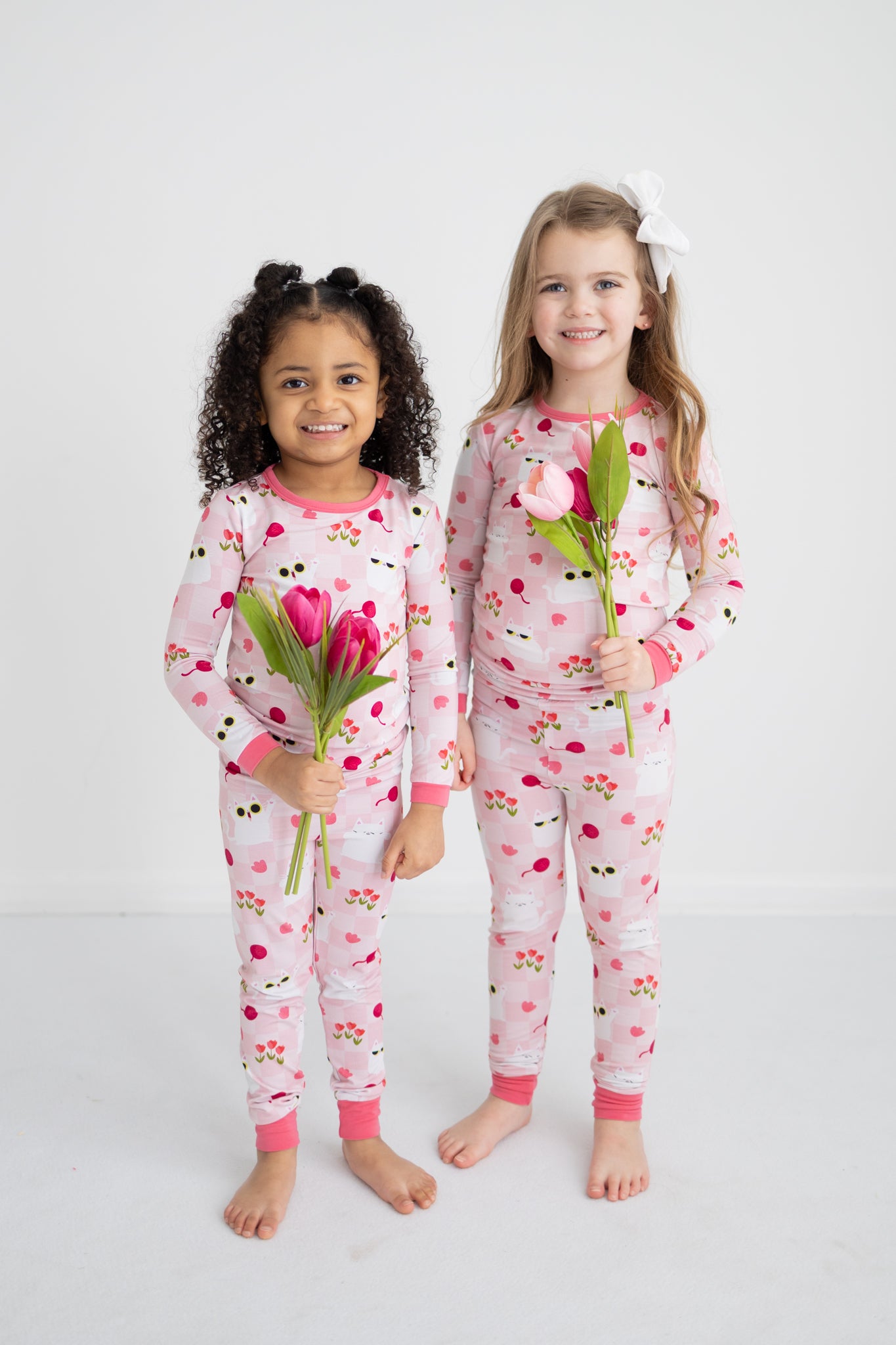 Two girls wearing matching pink pajamas with a cat and tulip print hold bouquets of tulips.