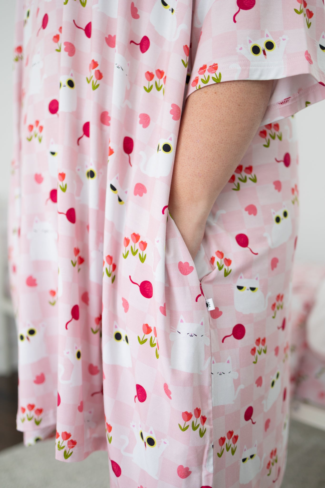 A person wears an oversized pink sleep tee featuring a white cat and tulip pattern with pockets.
