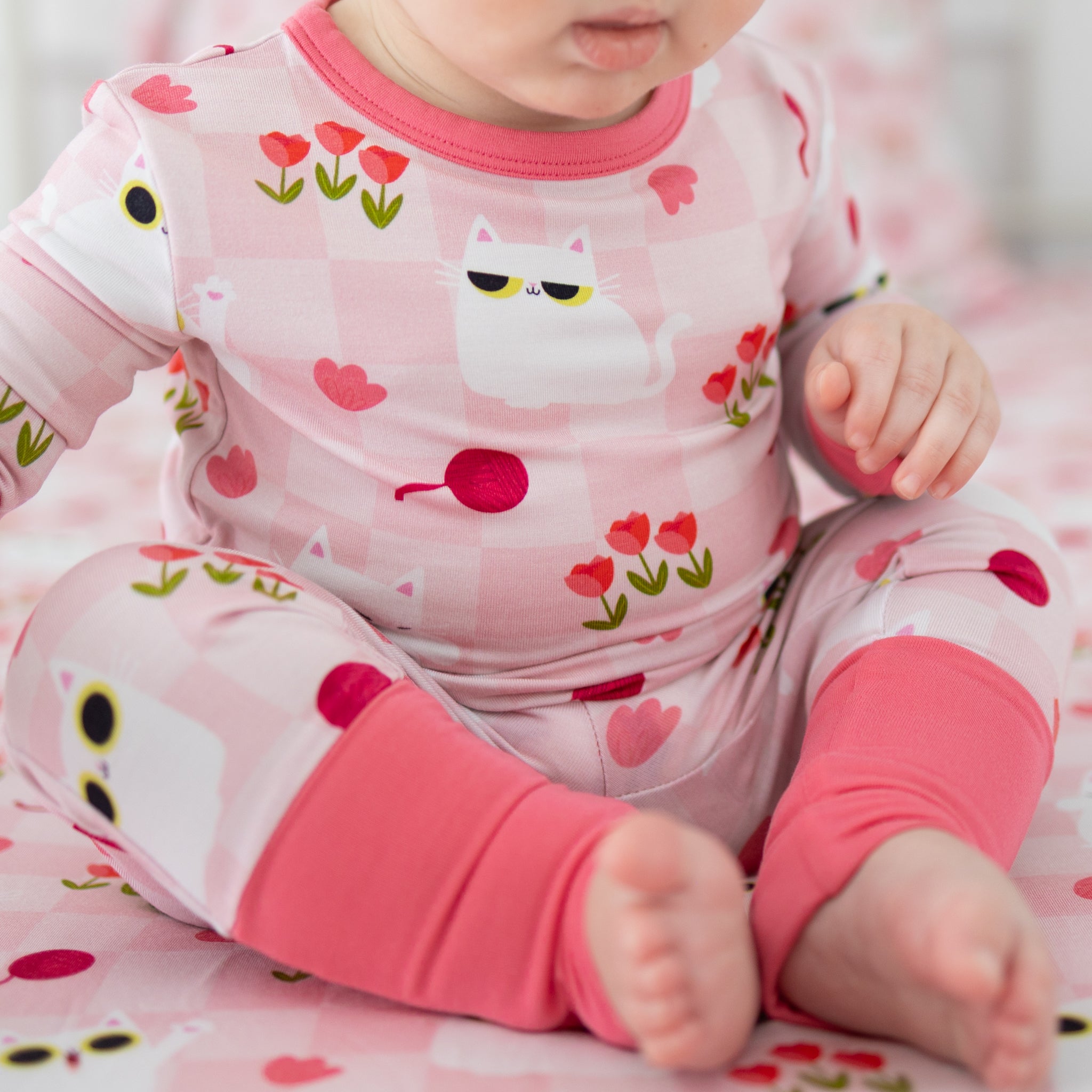 A baby wears a pink and white checkered pajama set featuring a cat and tulip print.
