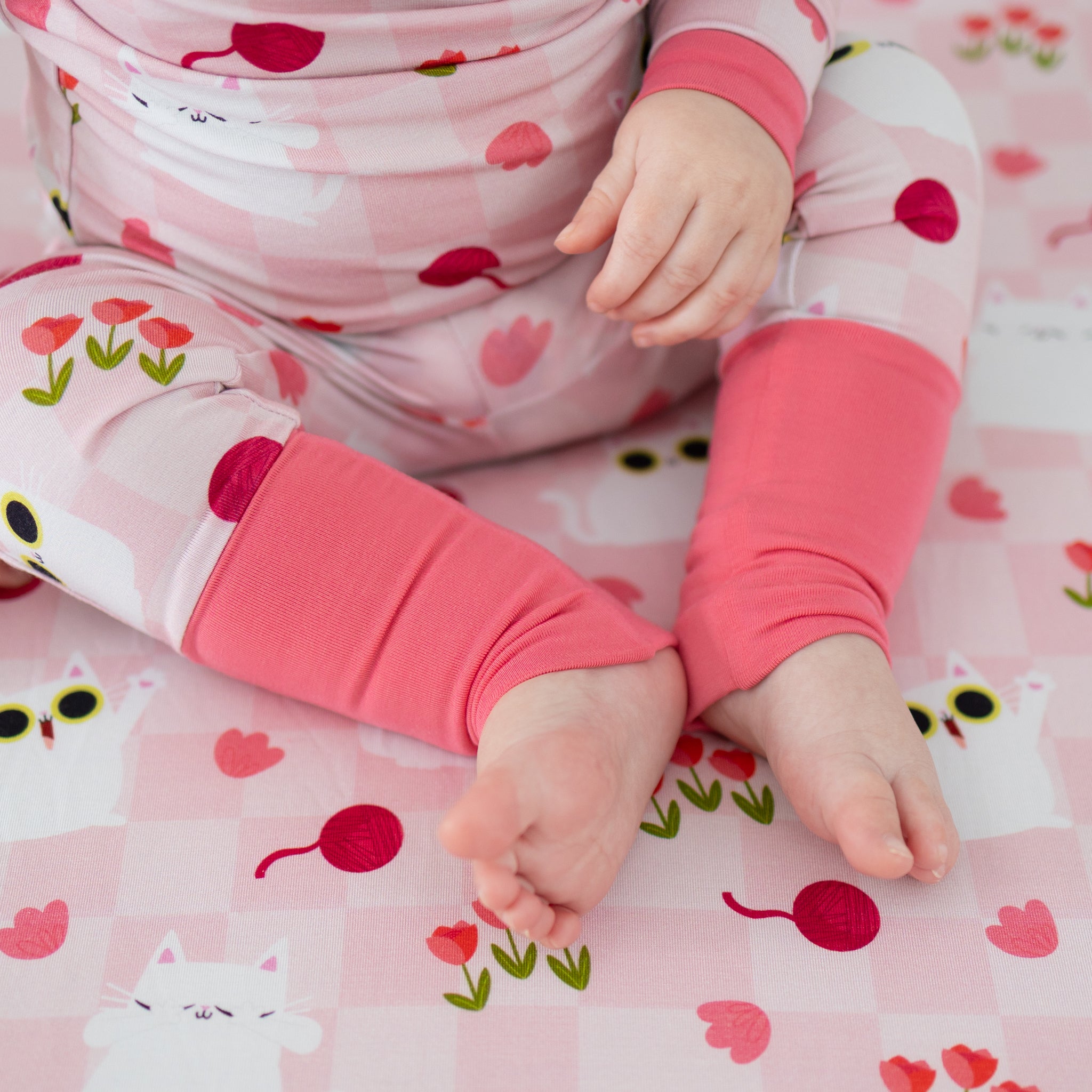 A baby wears pink pajamas with a white cat and tulip print featuring fold-over cuffs.