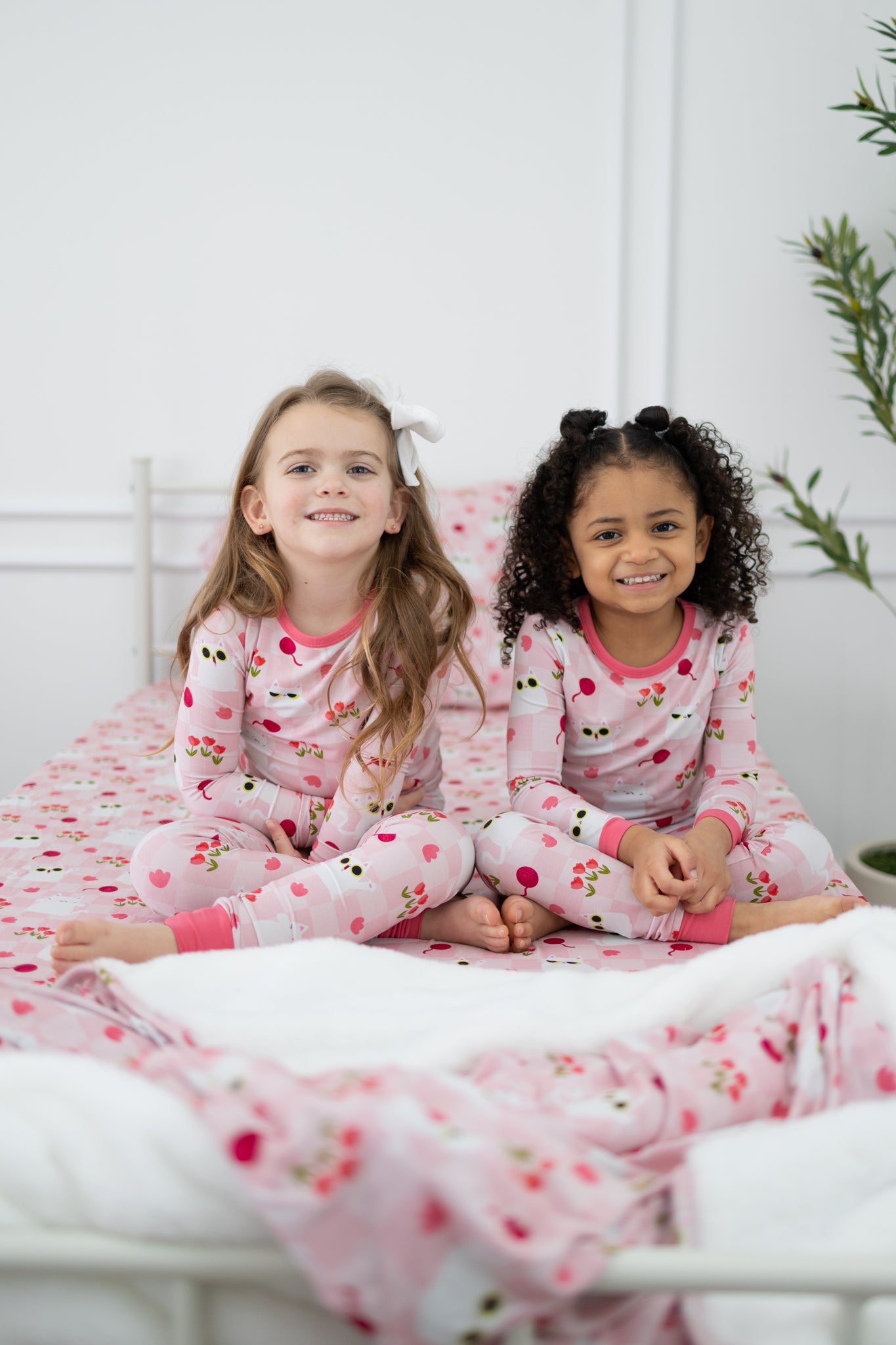 Two young girls sit on a bed wearing matching pink pajamas featuring a peek-a-boo cat print.