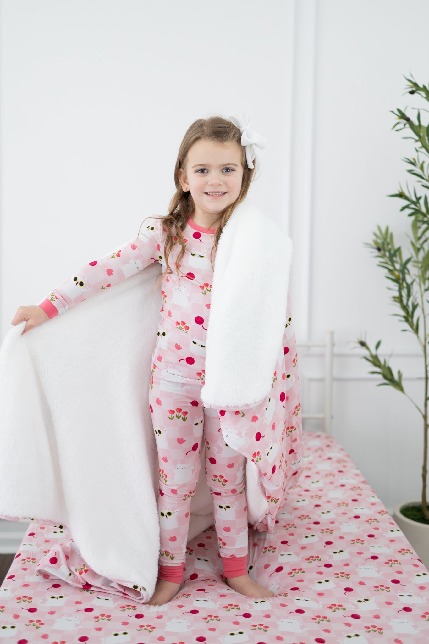 A child stands on a bed holding a reversible blanket with a white cat and pink floral pattern.
