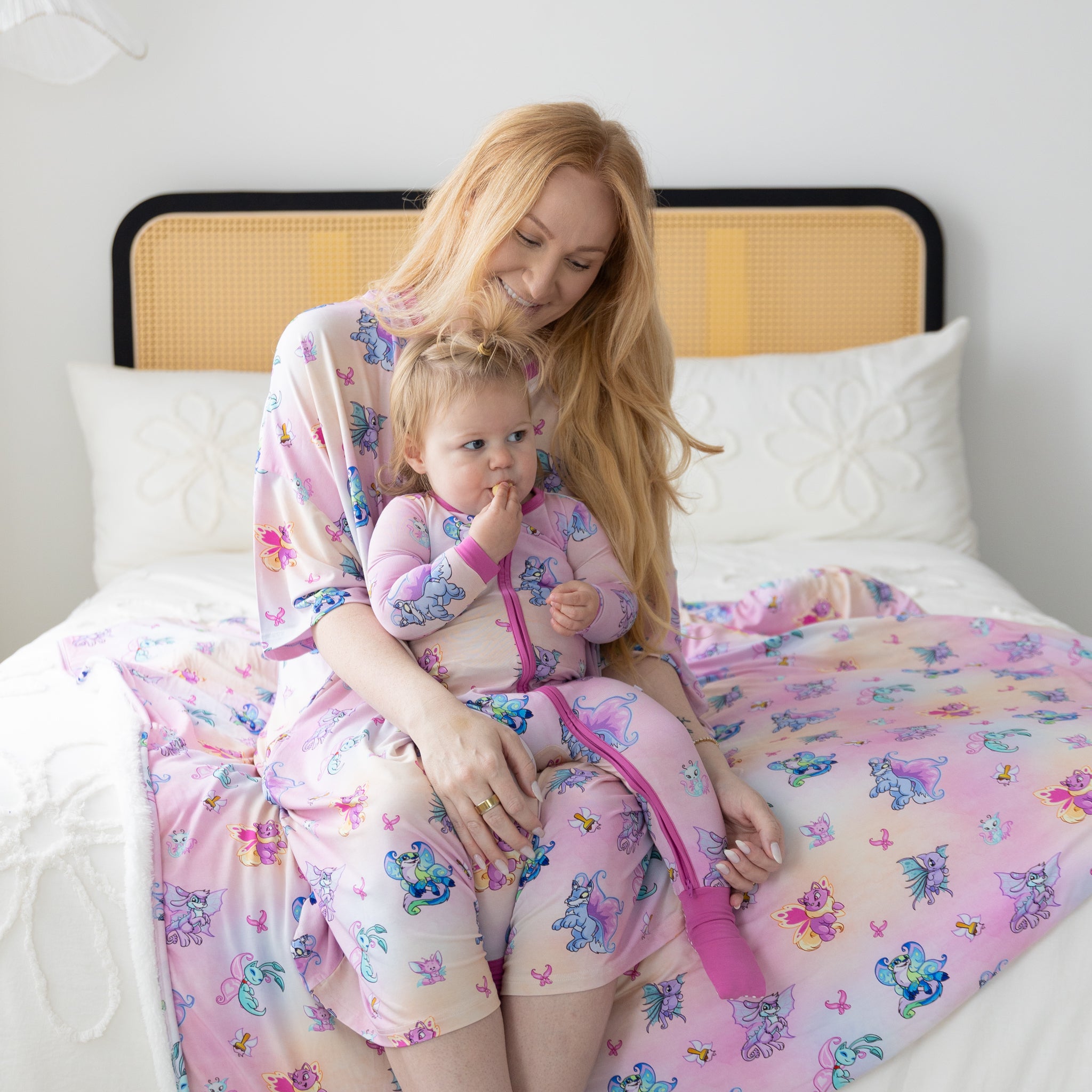 A mother and child sit on a bed wearing matching pink pajamas featuring Neopets Faerie prints.