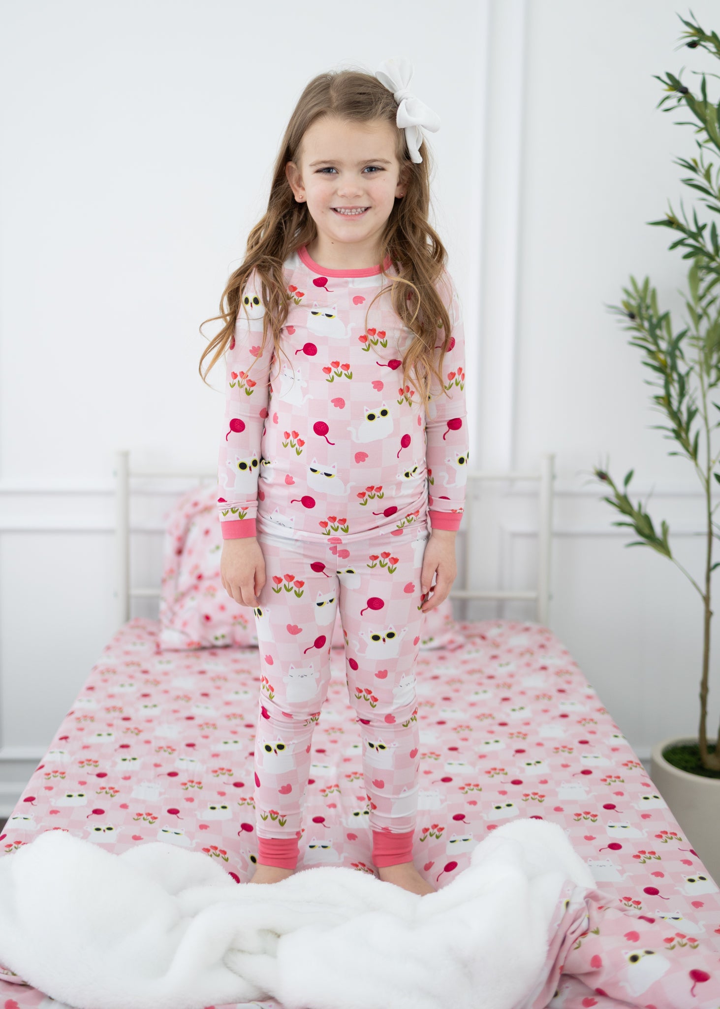 A young girl stands on a bed wearing a pink pajama set featuring a white cat and tulip pattern.