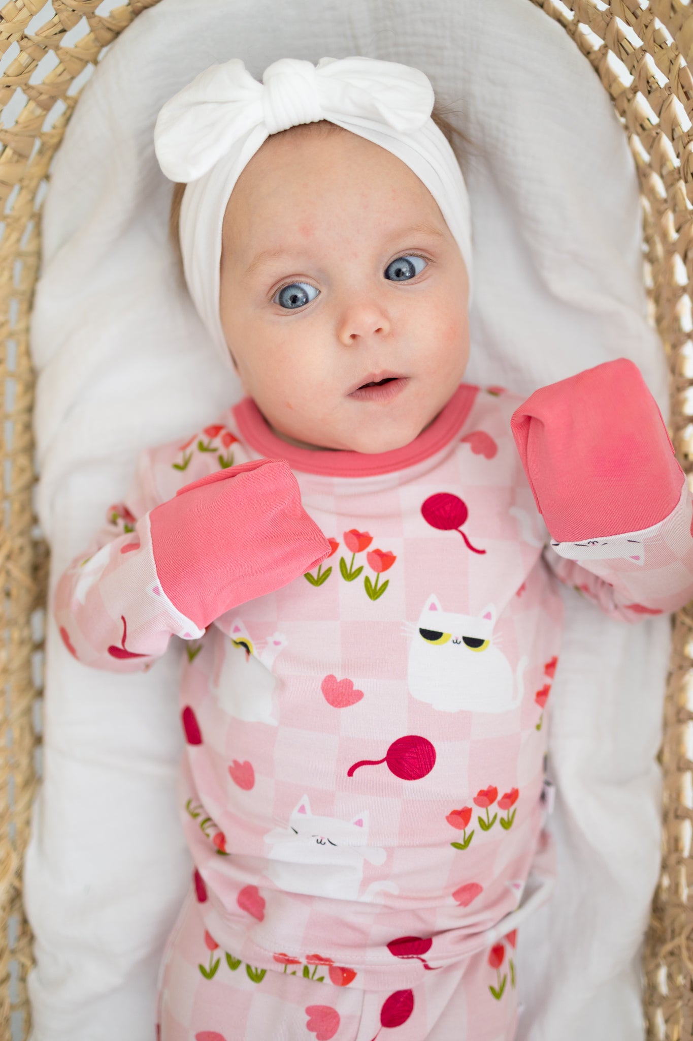 A baby wearing a pink cat-patterned pajama set with fold-over mitts and a white bow headband.