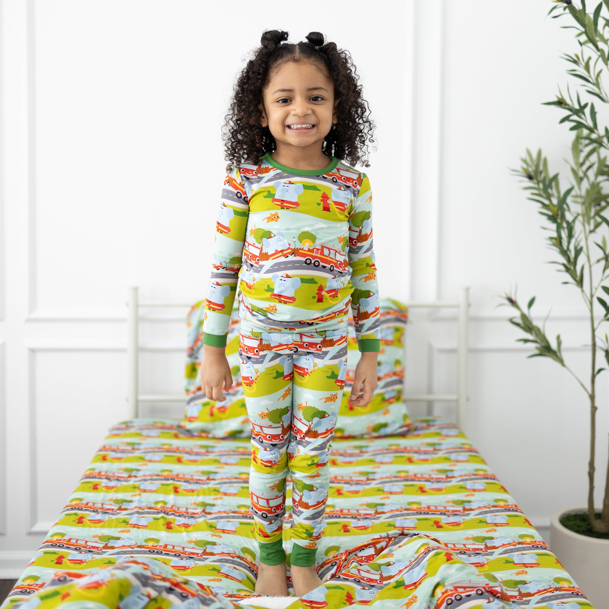 A child stands on a bed wearing a long-sleeve pajama set featuring a firetruck and animal print.