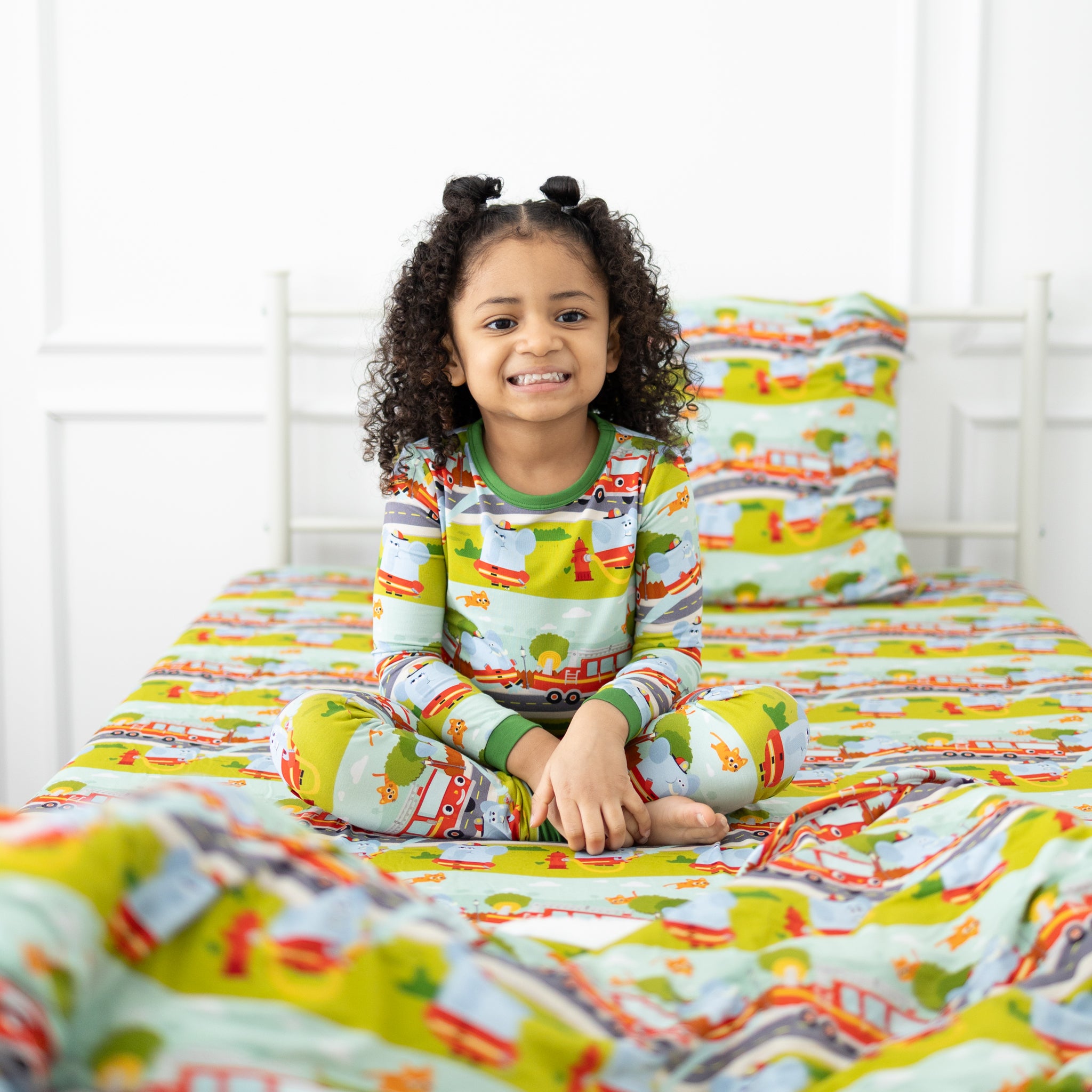 A child sits on a bed featuring a twin sheet and pillowcase set with a colorful firetruck pattern.