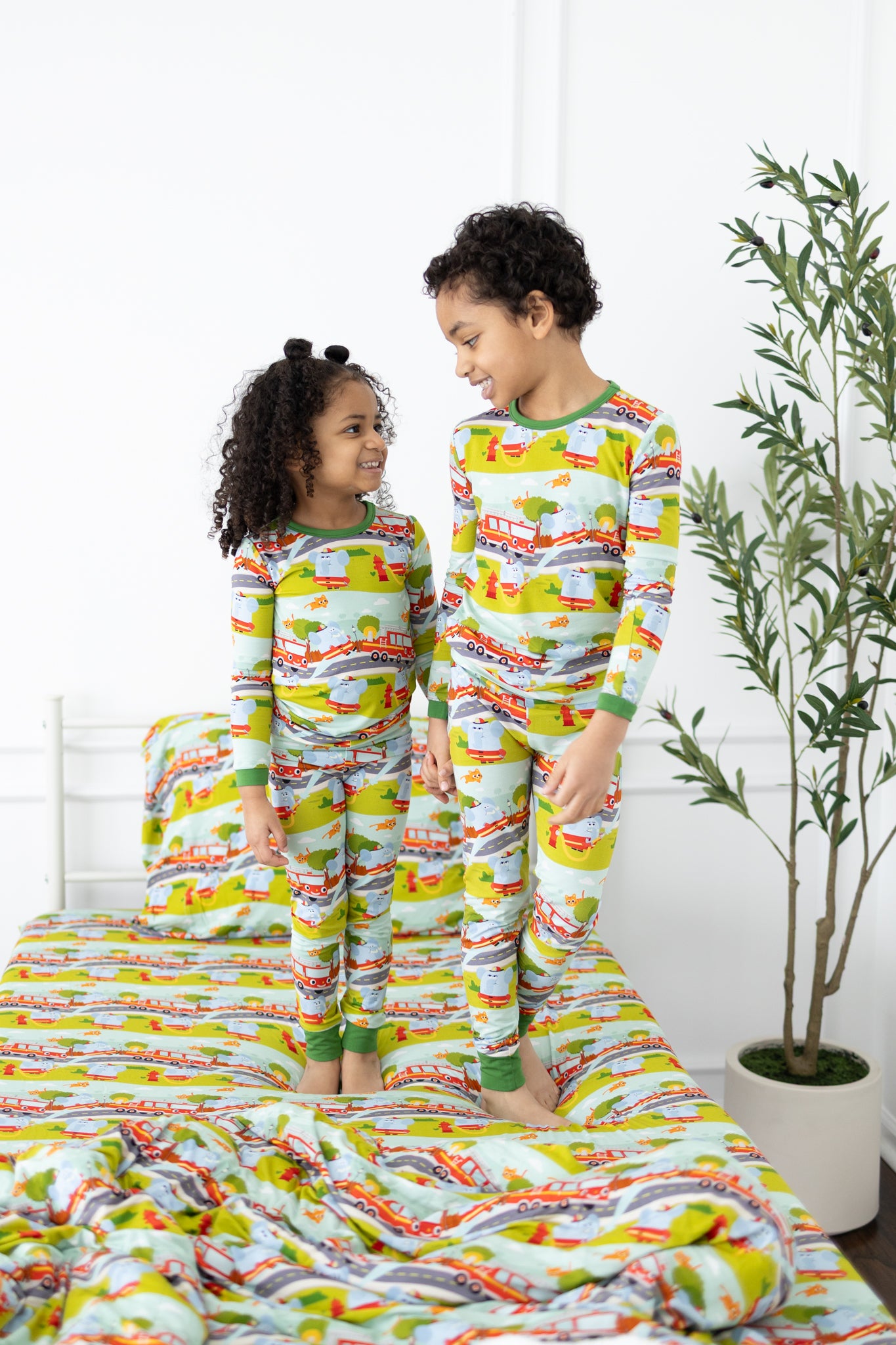 Two children stand on a bed wearing matching long-sleeve pajamas with a firetruck print.